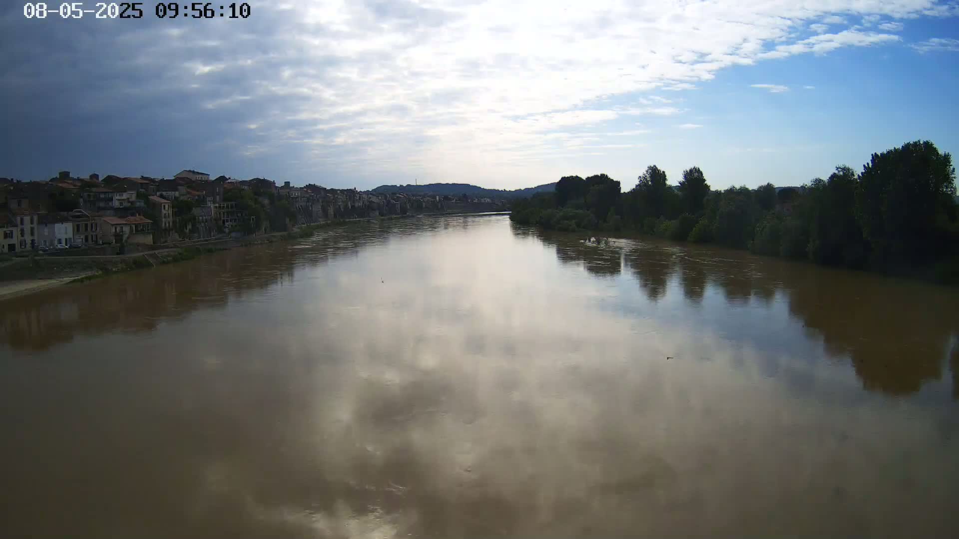 A wide, muddy river reflects a partly cloudy sky, with a town and trees lining its banks.