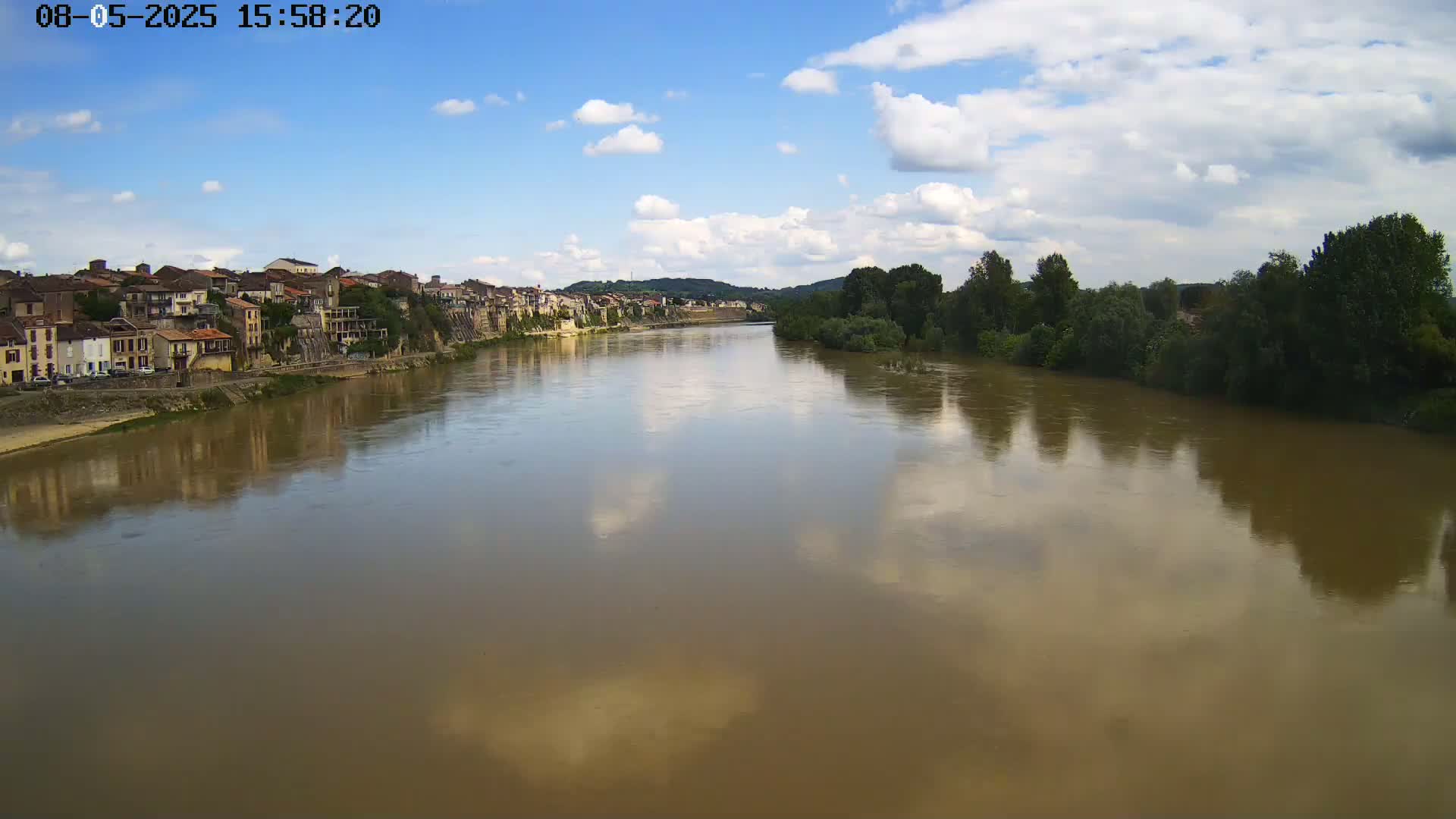 A wide, muddy river reflects a partly cloudy sky and the buildings of a town on its banks, lined with green trees.