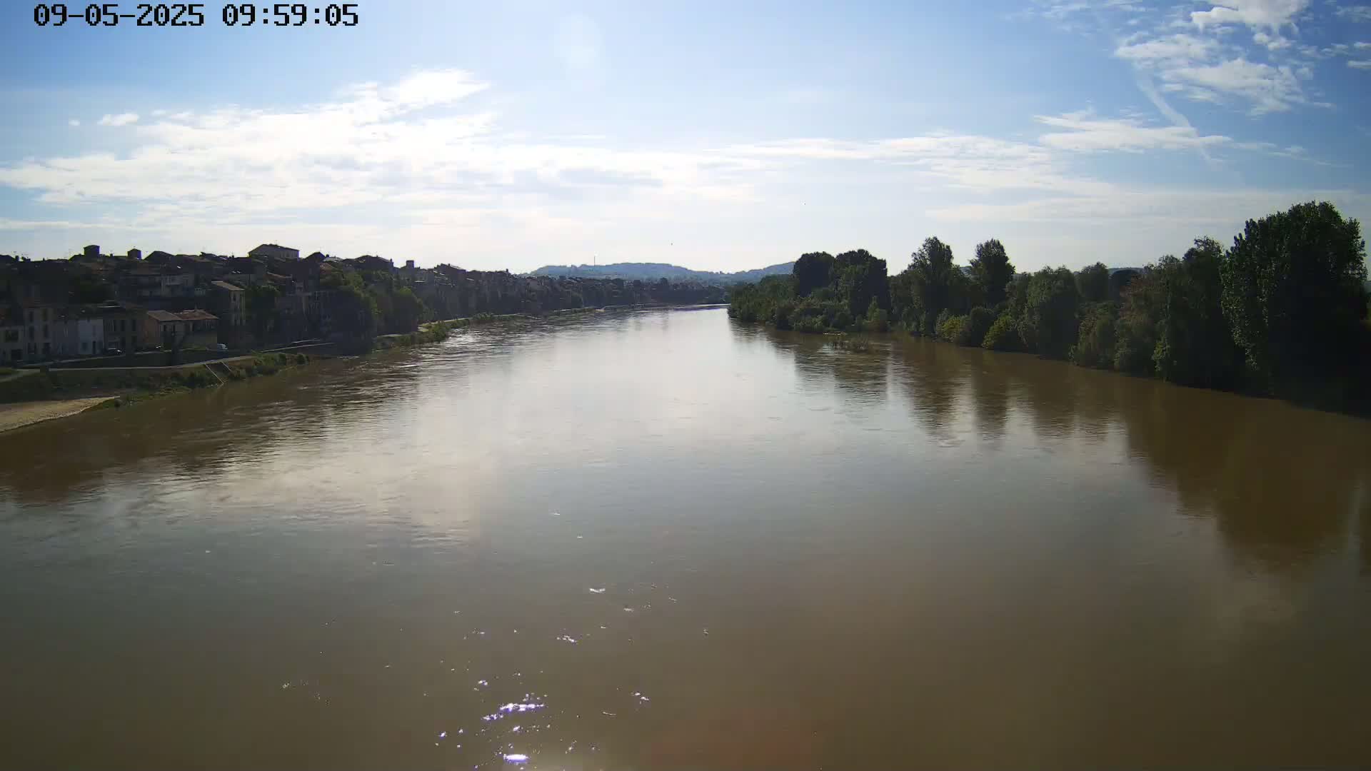 A wide, brown river flows under a partly cloudy sky past a town on one bank and trees on the other.
