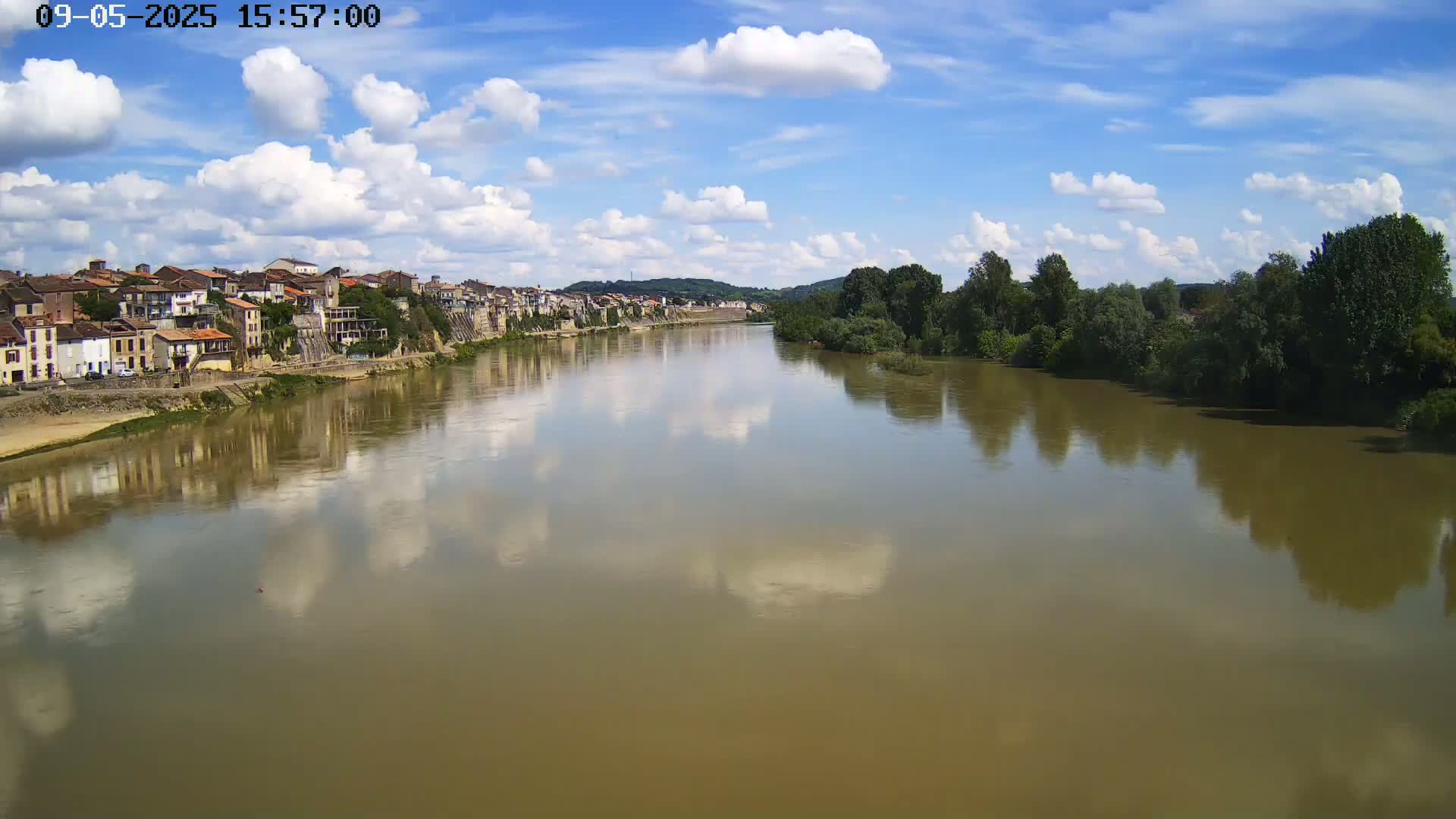A wide, calm river reflects a partly cloudy blue sky and the buildings of a town on its bank, lined with green trees on the opposite shore.