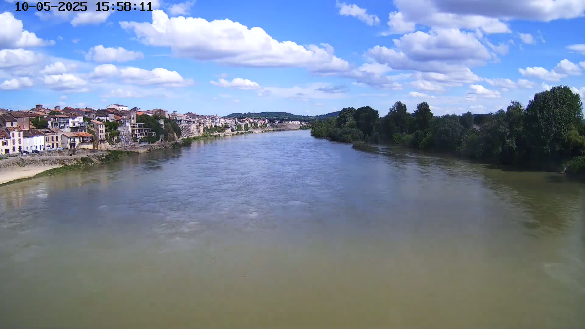 A wide, murky river flows under a partly cloudy blue sky past a riverside town with buildings of varying heights and colors.