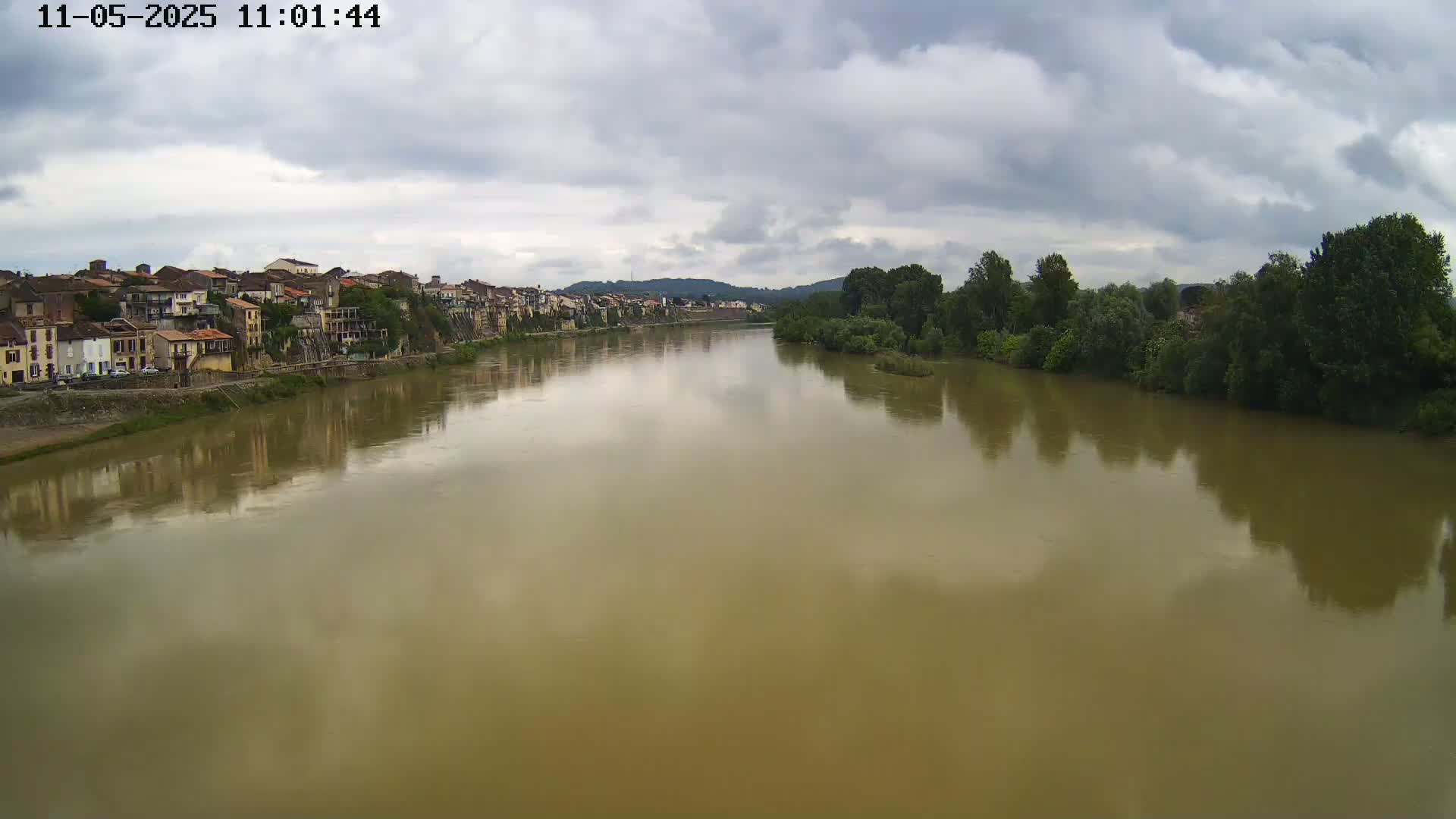 A wide, muddy river reflects a cloudy sky and a line of buildings on one bank, with green trees lining the opposite bank.