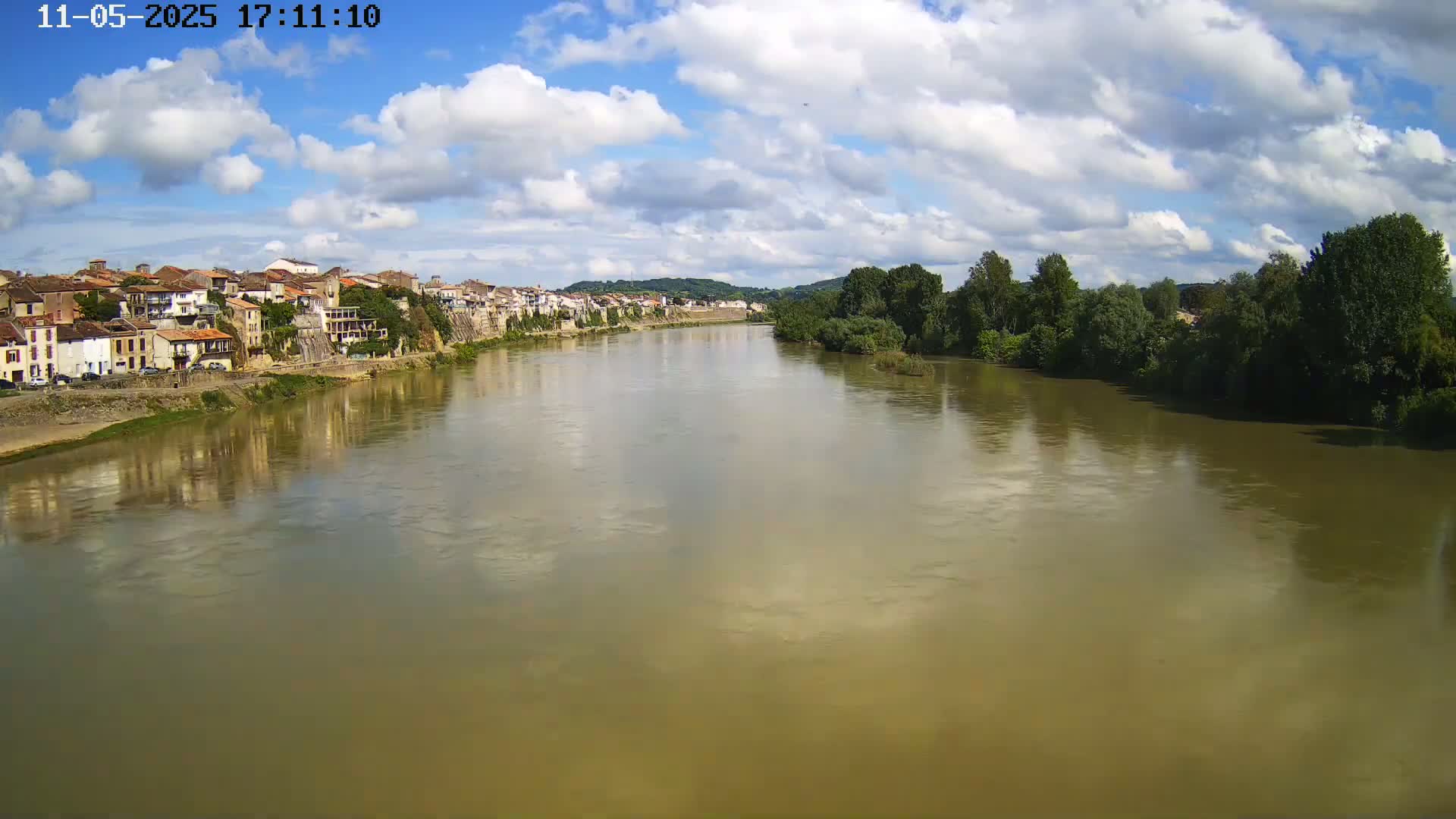 A wide, muddy river reflects a partly cloudy sky, with a town of buildings lining one bank and green trees along the other.