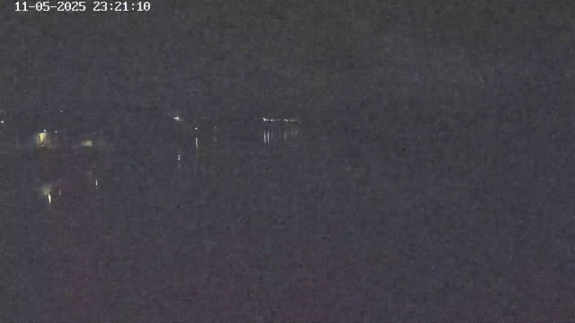 A nighttime, dark, grainy image shows a body of water reflecting faint lights from a distant shoreline.