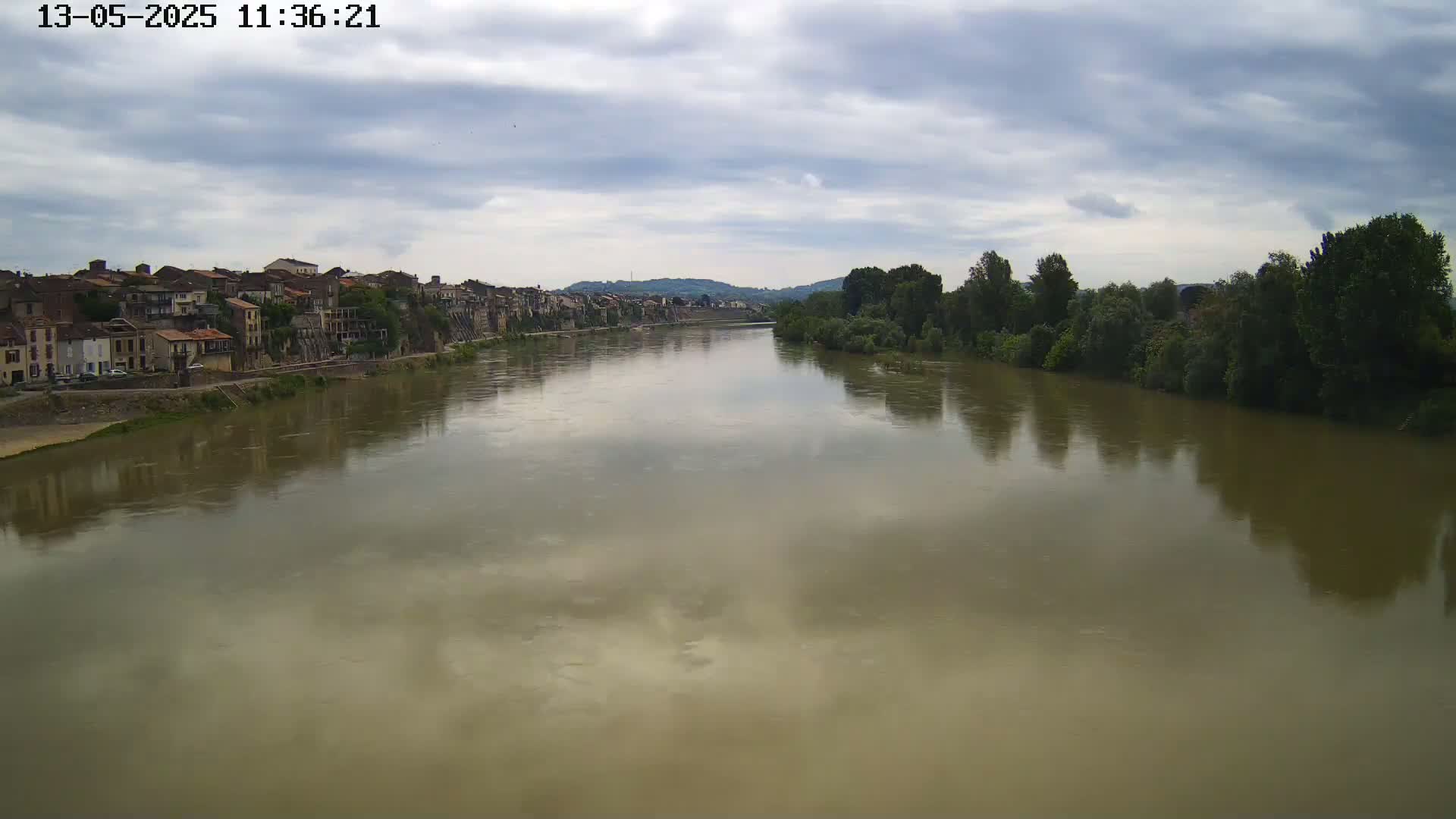 A wide, muddy river reflects a cloudy sky, with a town visible on one bank and trees lining the opposite shore.