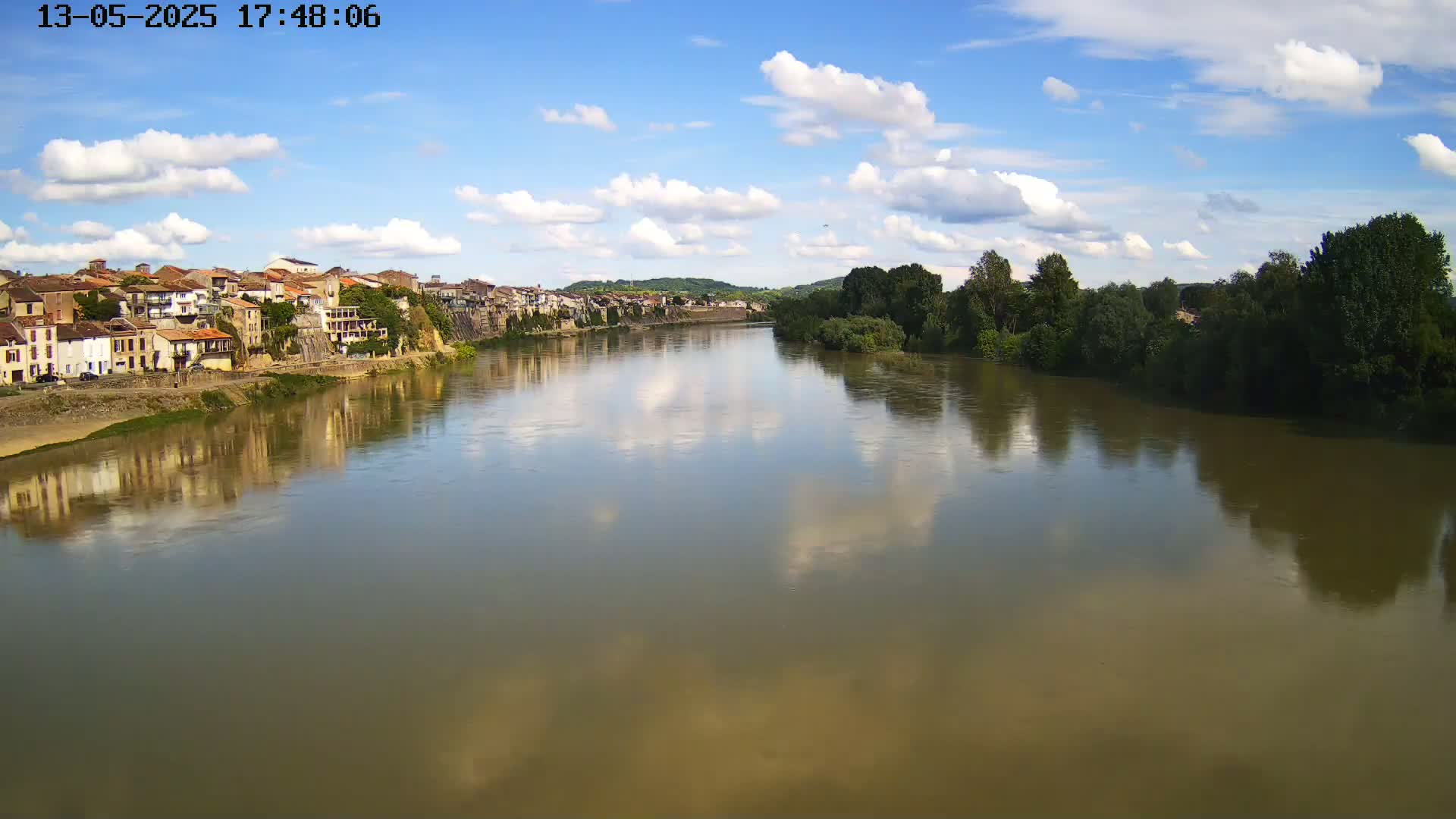 A river reflects a partly cloudy sky and the buildings of a town on its bank.