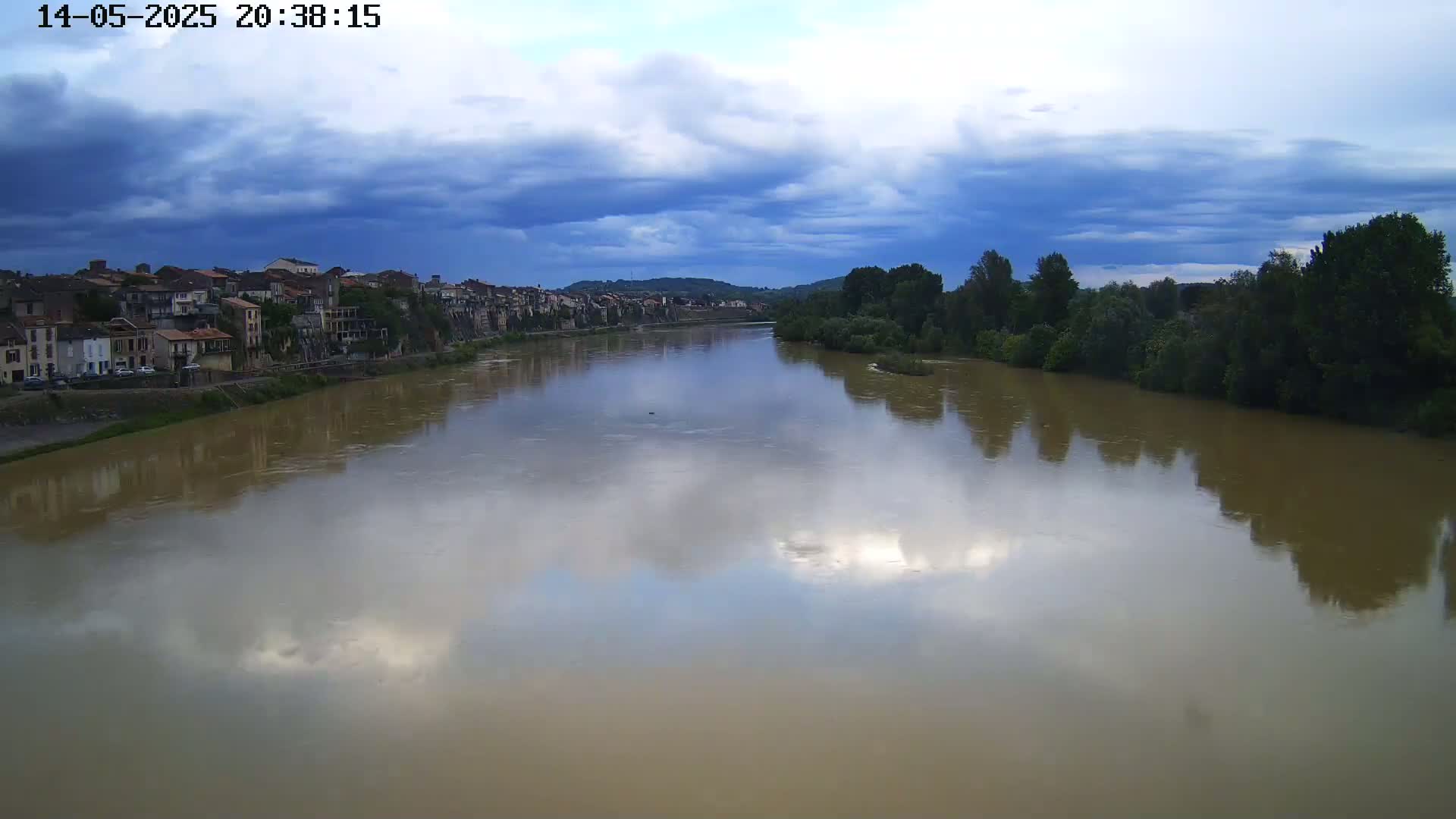 A wide, muddy river reflects a partly cloudy sky under which a town sits on its bank, lined with trees on the opposite shore.