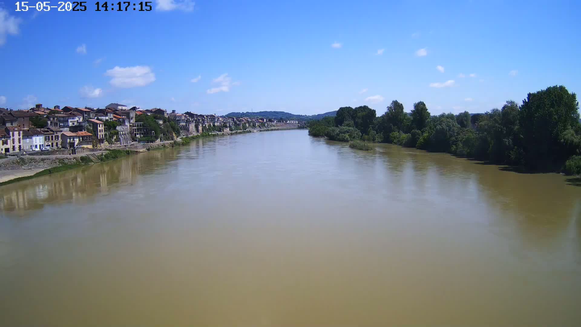 A wide, muddy river flows under a mostly sunny sky past a town of closely-packed buildings and lush green trees.