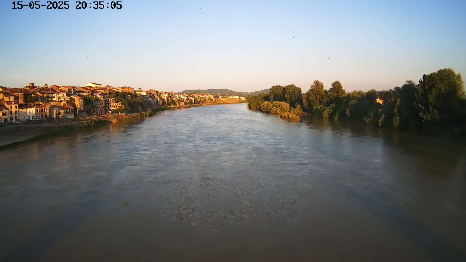 A wide, calm river flows past a town of low, tan buildings and lush green trees under a clear, dusky sky.