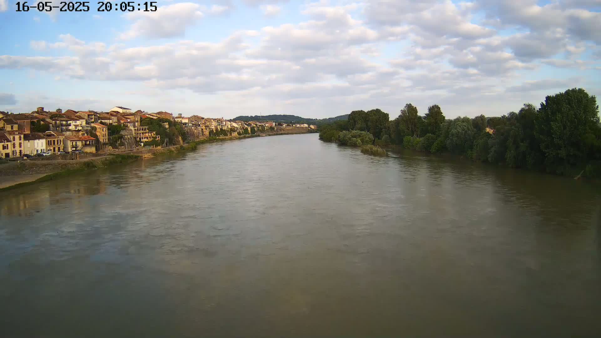 A wide, calm river flows past a town of buildings nestled along its bank, under a partly cloudy sky.