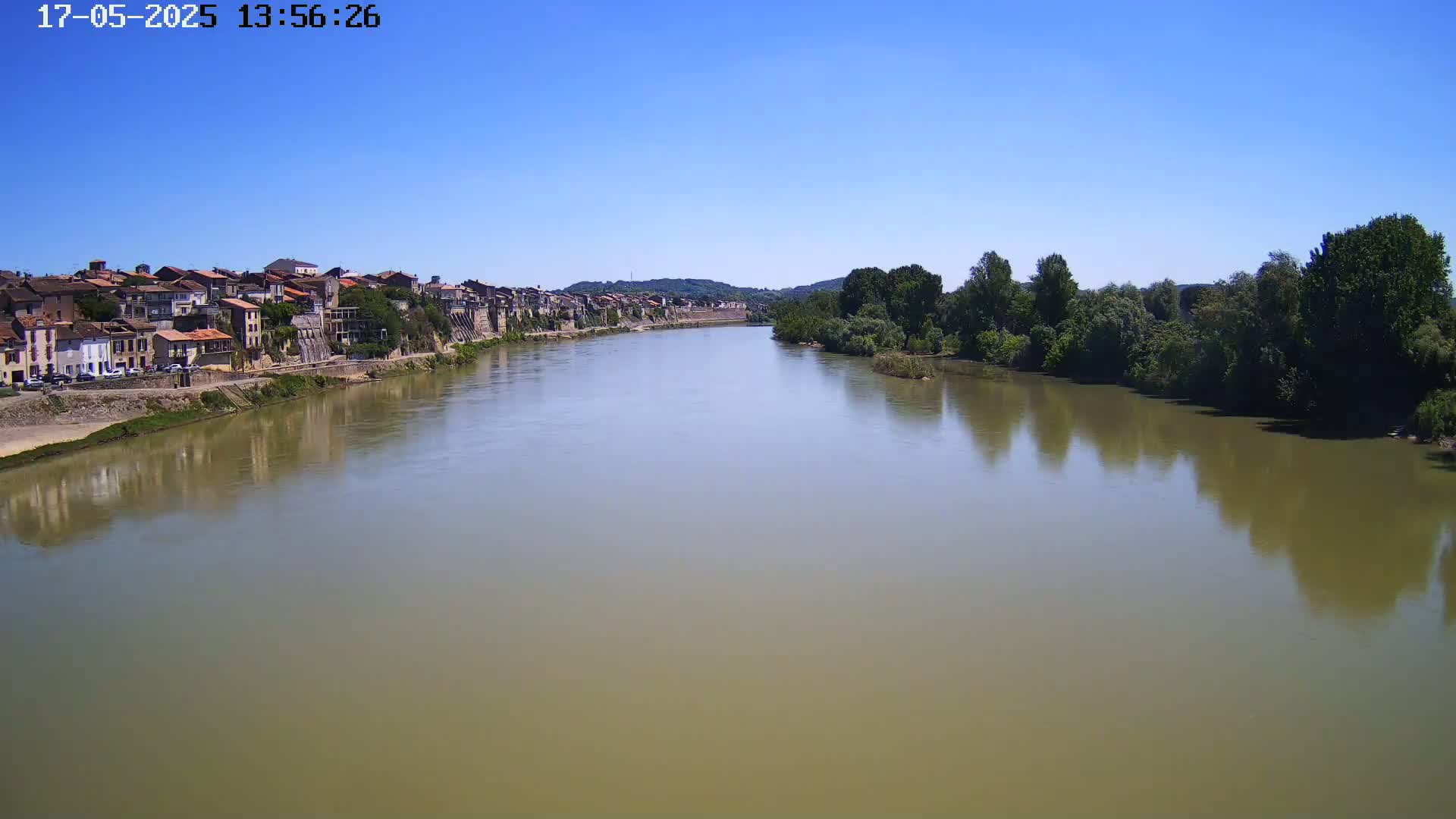 A wide, calm river with a light brown tint flows past a town with buildings along the bank under a clear blue sky.