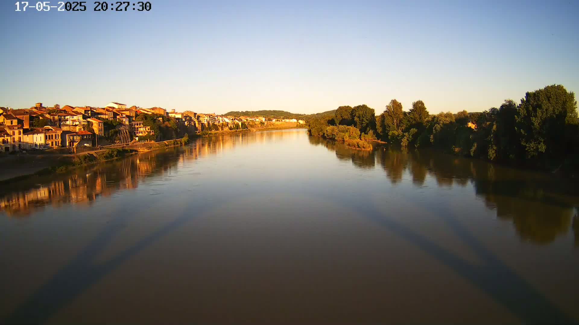 A calm river reflects the setting sun and the shadows of a bridge on its surface, with a town visible on the opposite bank and trees lining the river's edge.