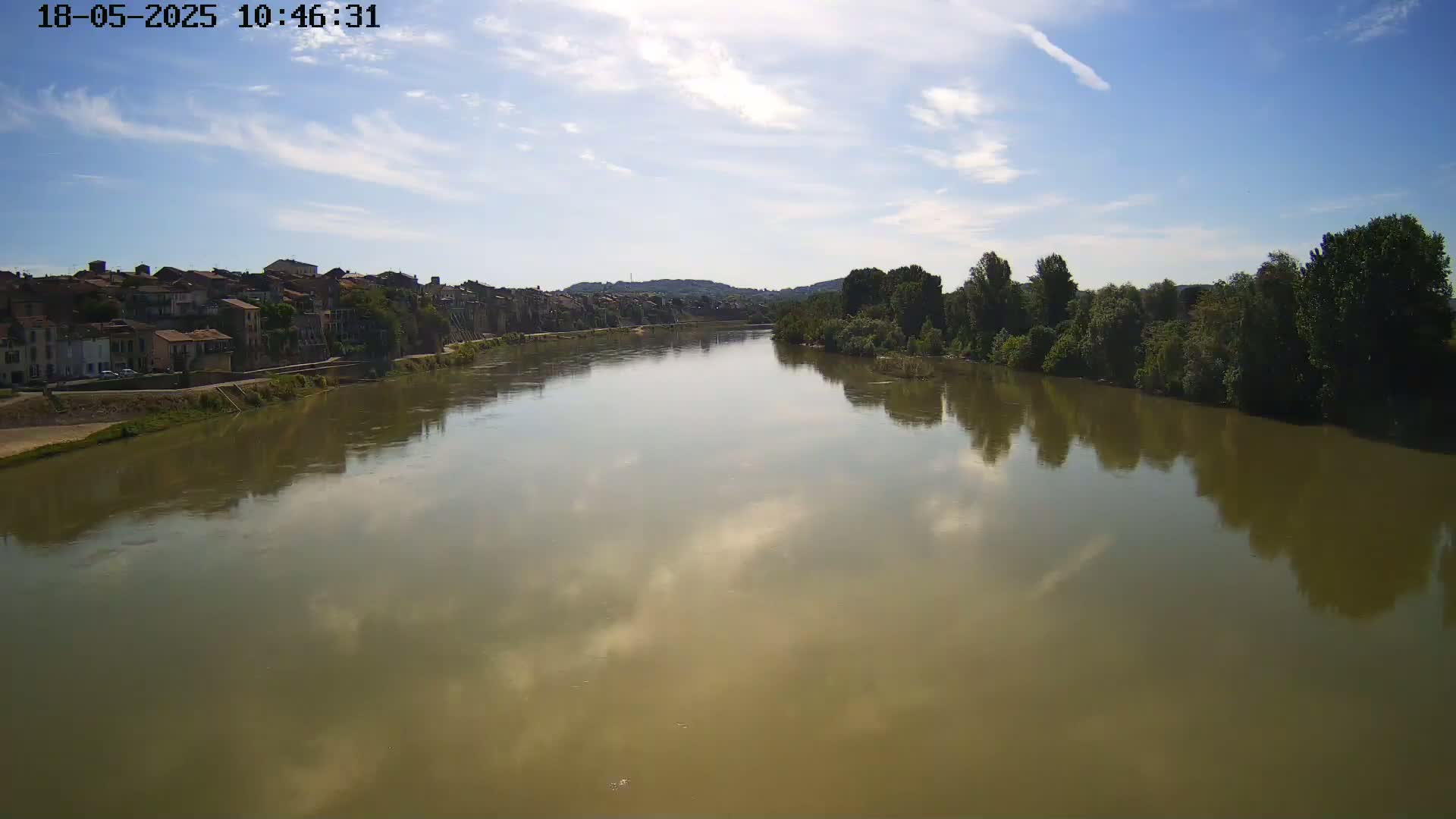 A wide, calm river with a muddy-brown color reflects a partly cloudy sky, with a town and trees visible on its banks.
