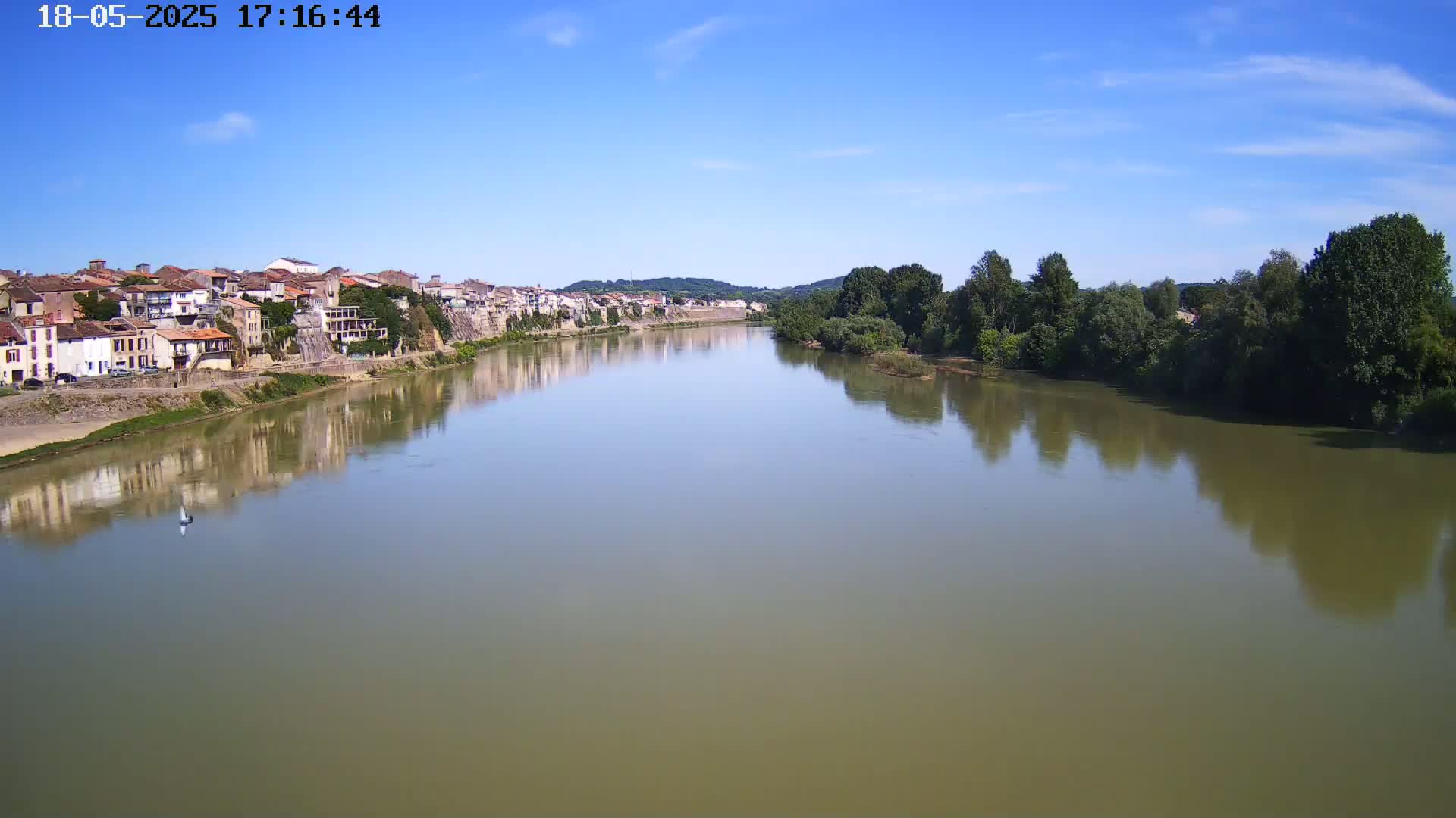 A calm river reflects a town and green trees under a clear blue sky.
