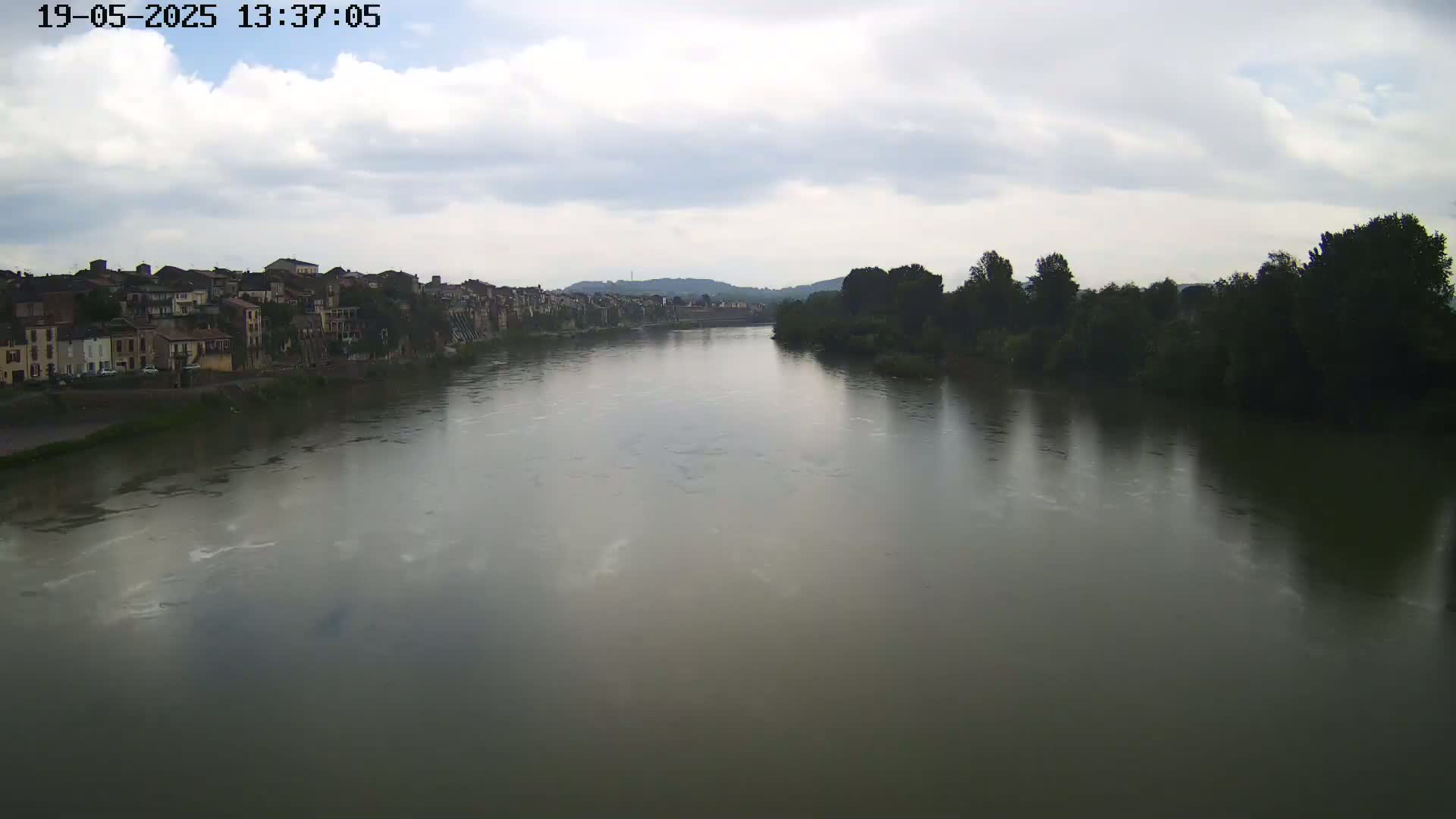 A wide river flows under a cloudy sky past a town on one bank and trees on the other.