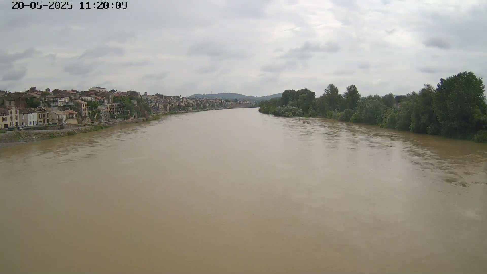 A wide, muddy river flows under an overcast sky past a line of buildings and trees.