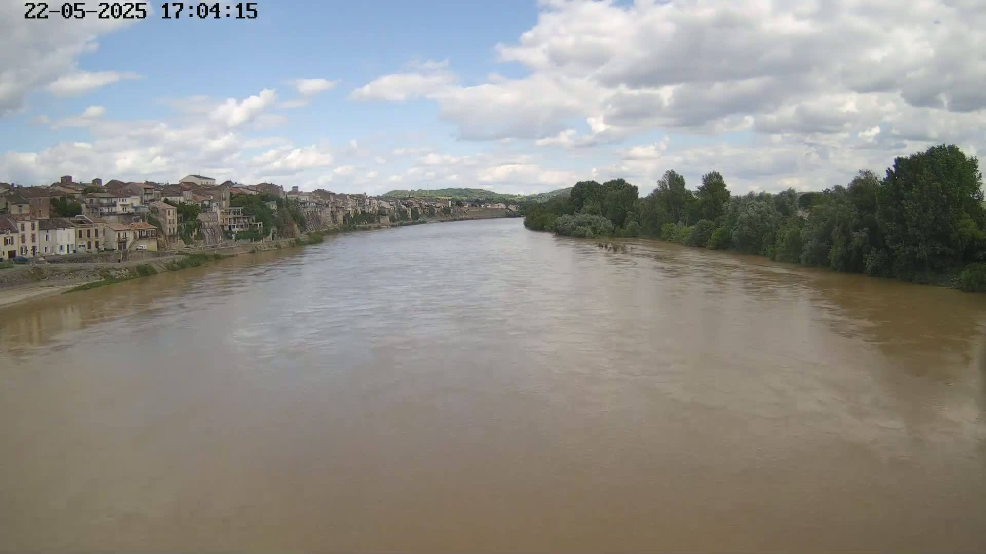 A wide, muddy river flows under a partly cloudy sky past a town built on a hillside.