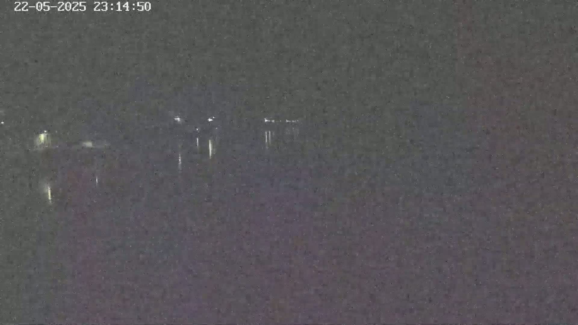 A nighttime, low-light image shows a dark body of water with faint lights reflecting on its surface from a distant shoreline.