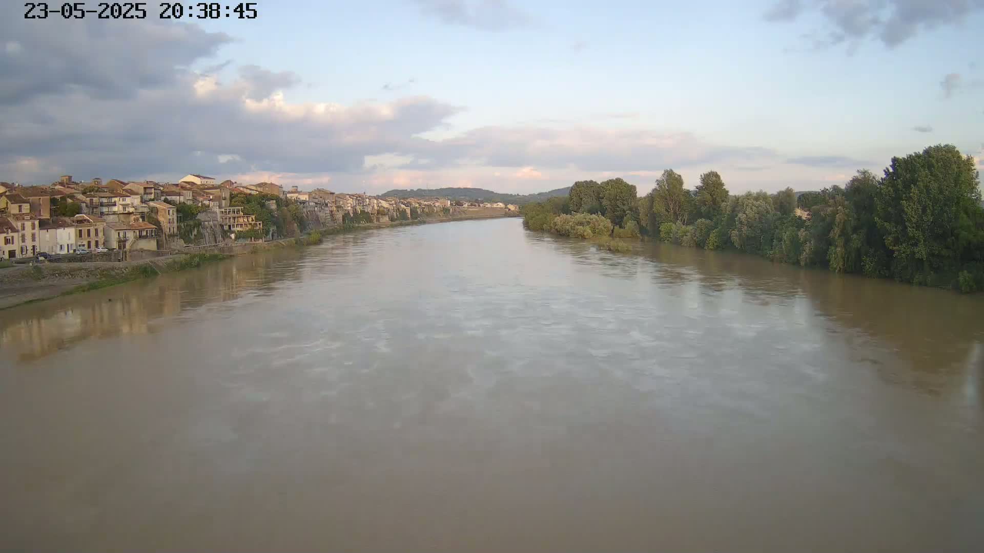 A wide, muddy river flows past a town under a partly cloudy sky.