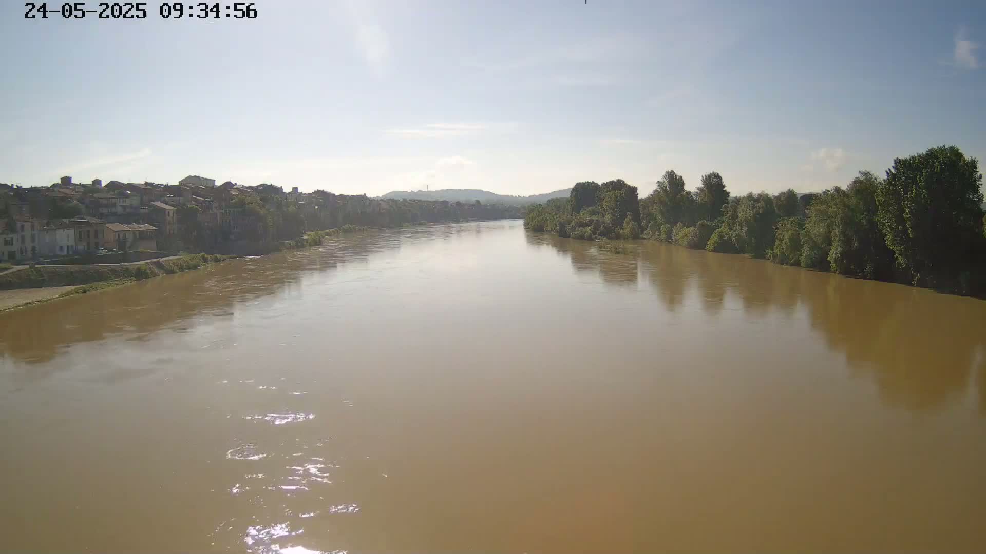 A wide, muddy river flows under a clear sky past a town and leafy riverbanks.