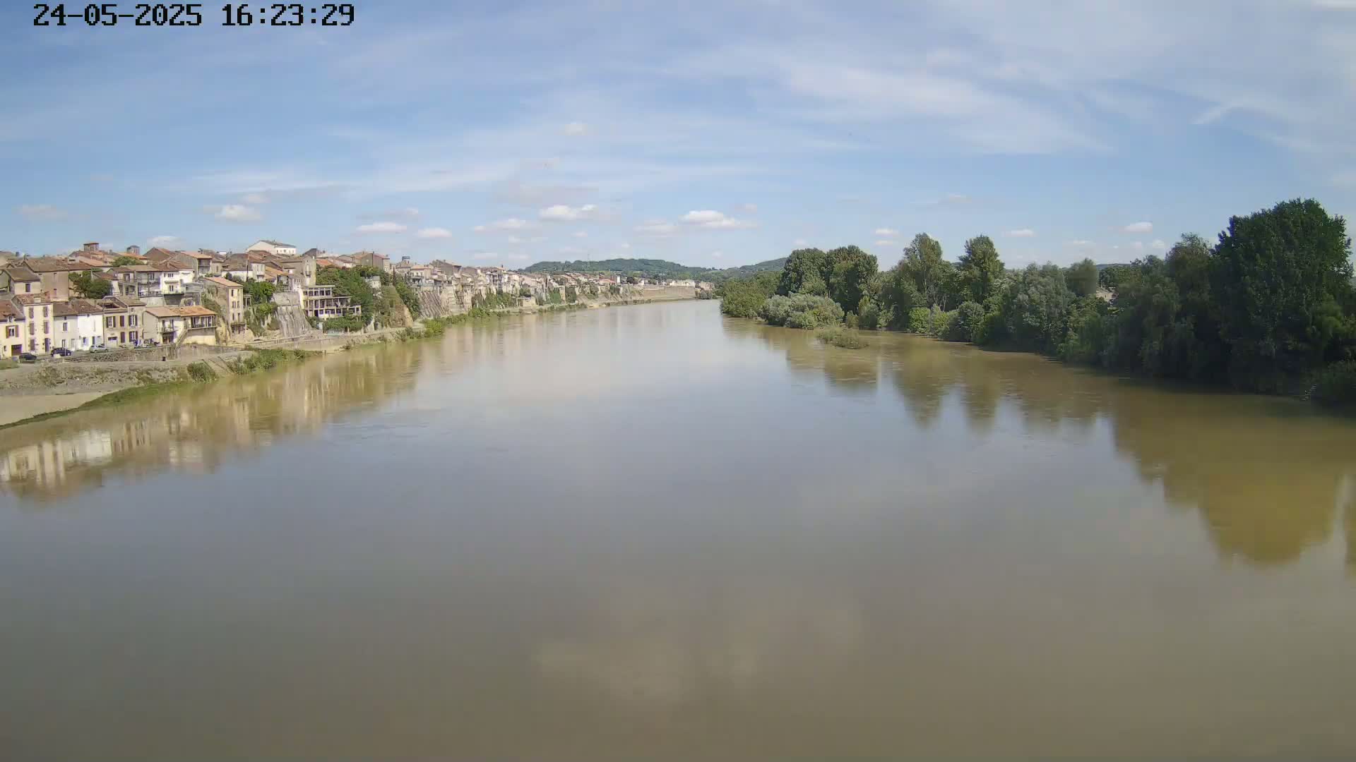A wide, muddy river flows under a partly cloudy sky, past a town of closely clustered buildings on its bank and a line of green trees on the opposite bank.