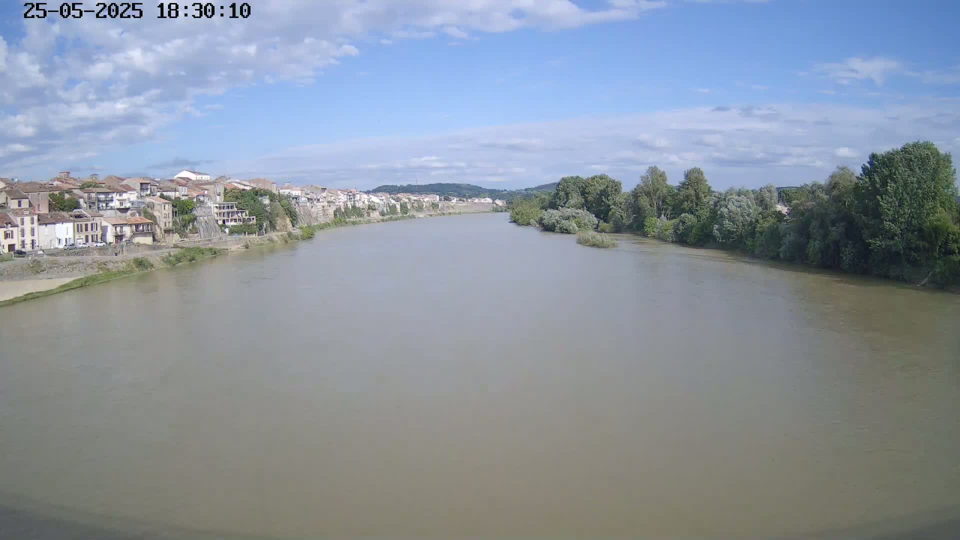 A wide, muddy river flows under a partly cloudy sky, past a town with buildings lining one bank and green trees along the other.