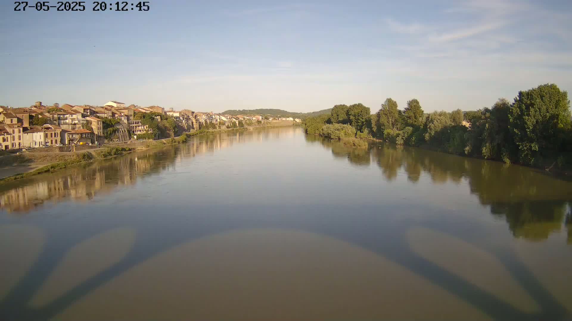 A calm river reflects a town and trees under a clear, bright sky.