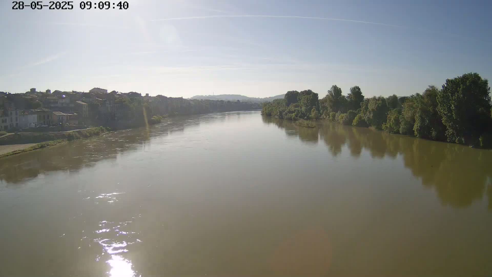 A wide, murky river flows under a clear, sunny sky past a town and tree-lined banks.