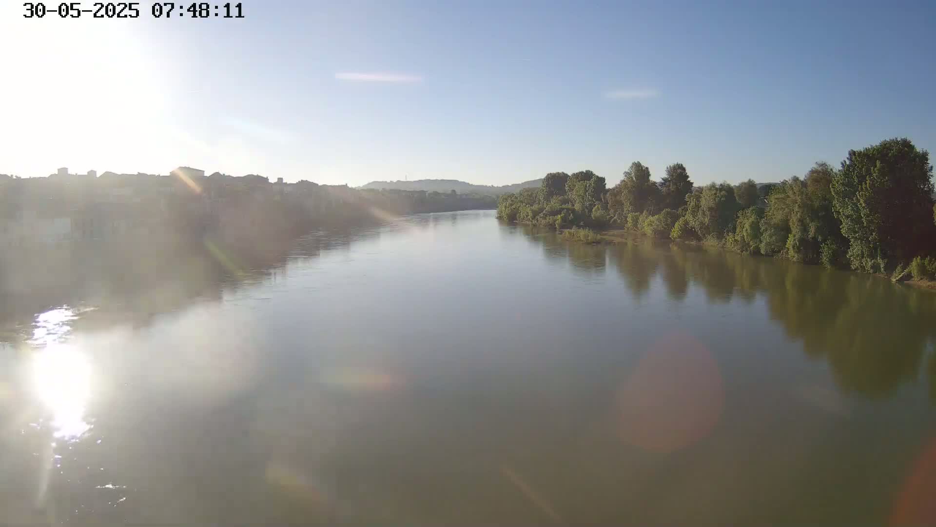 A wide, calm river reflects the sunlight on a clear, sunny day, flowing past a town on one bank and lush green trees on the other.