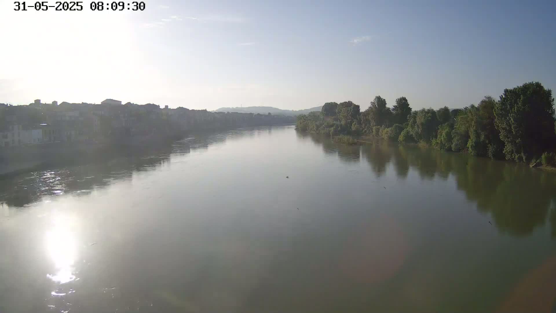 A wide, calm river reflects a hazy sunrise, with a town visible on one bank and lush greenery on the other.