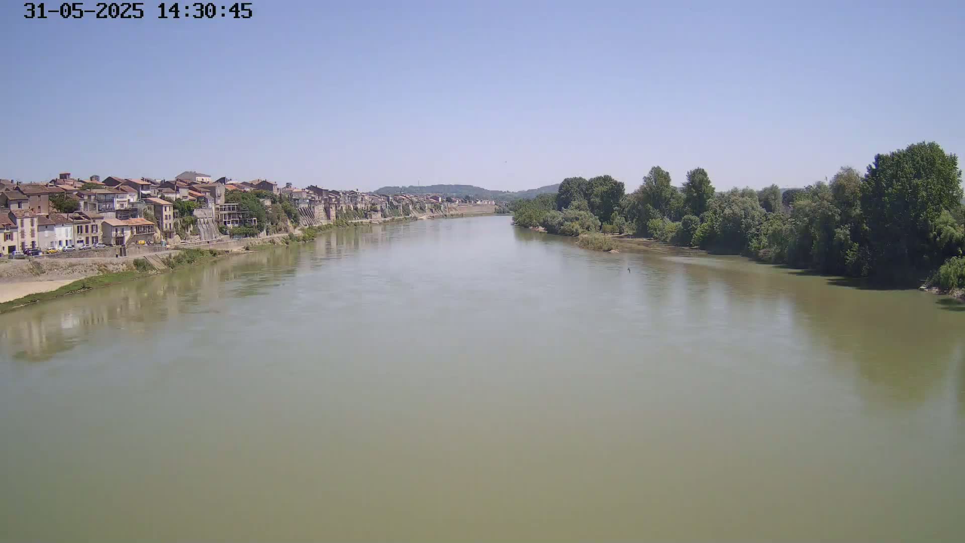 A wide, muddy river flows past a town of closely packed buildings on one bank and lush green trees on the other, under a clear, sunny sky.