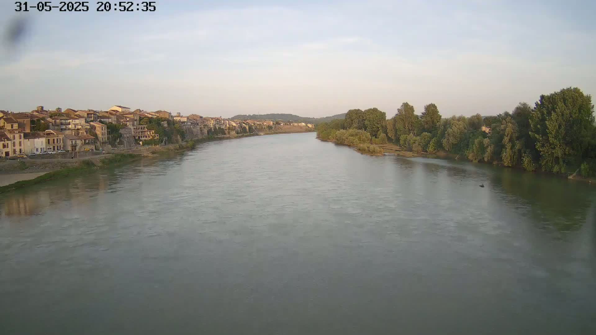 A wide, calm river flows past a town of closely-packed buildings under a pale, clear sky.