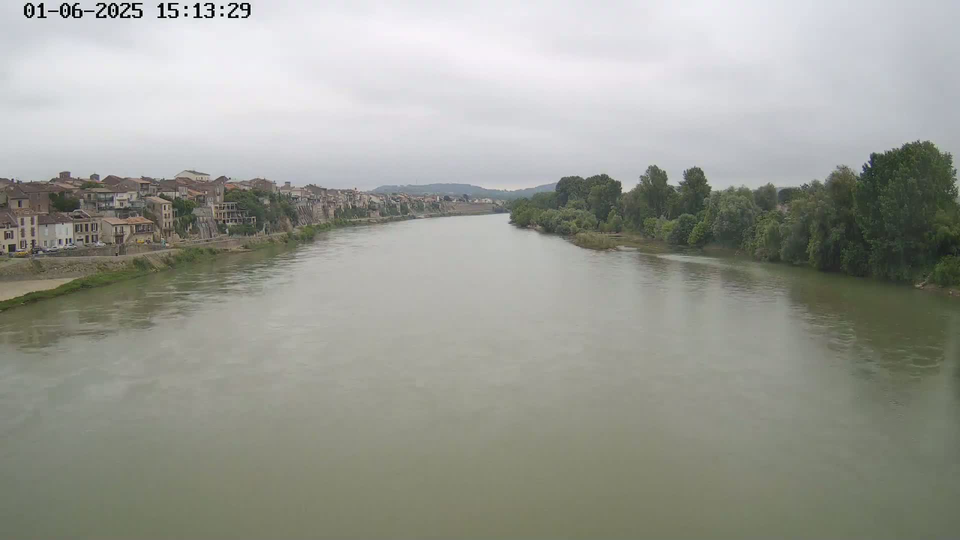 A wide, calm river flows past a town of closely-packed buildings under an overcast sky.