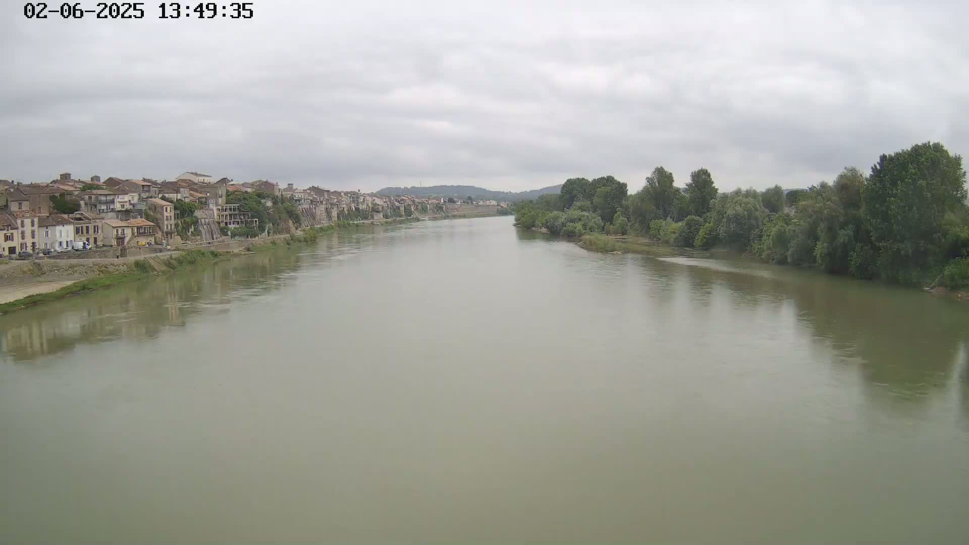 A wide, calm river flows under an overcast sky past a town situated on its bank, lined with green vegetation on both sides.
