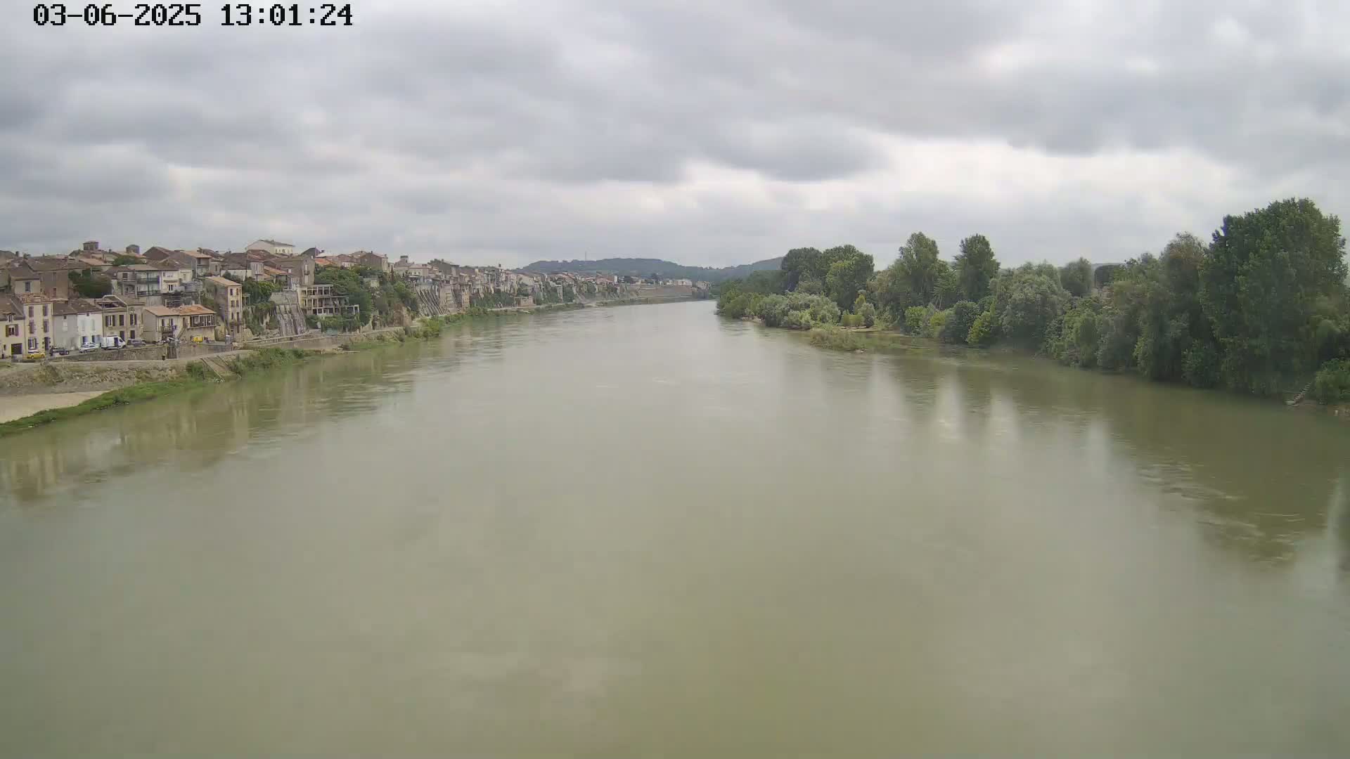 A wide, murky river flows under a cloudy sky past a line of buildings and trees on either bank.