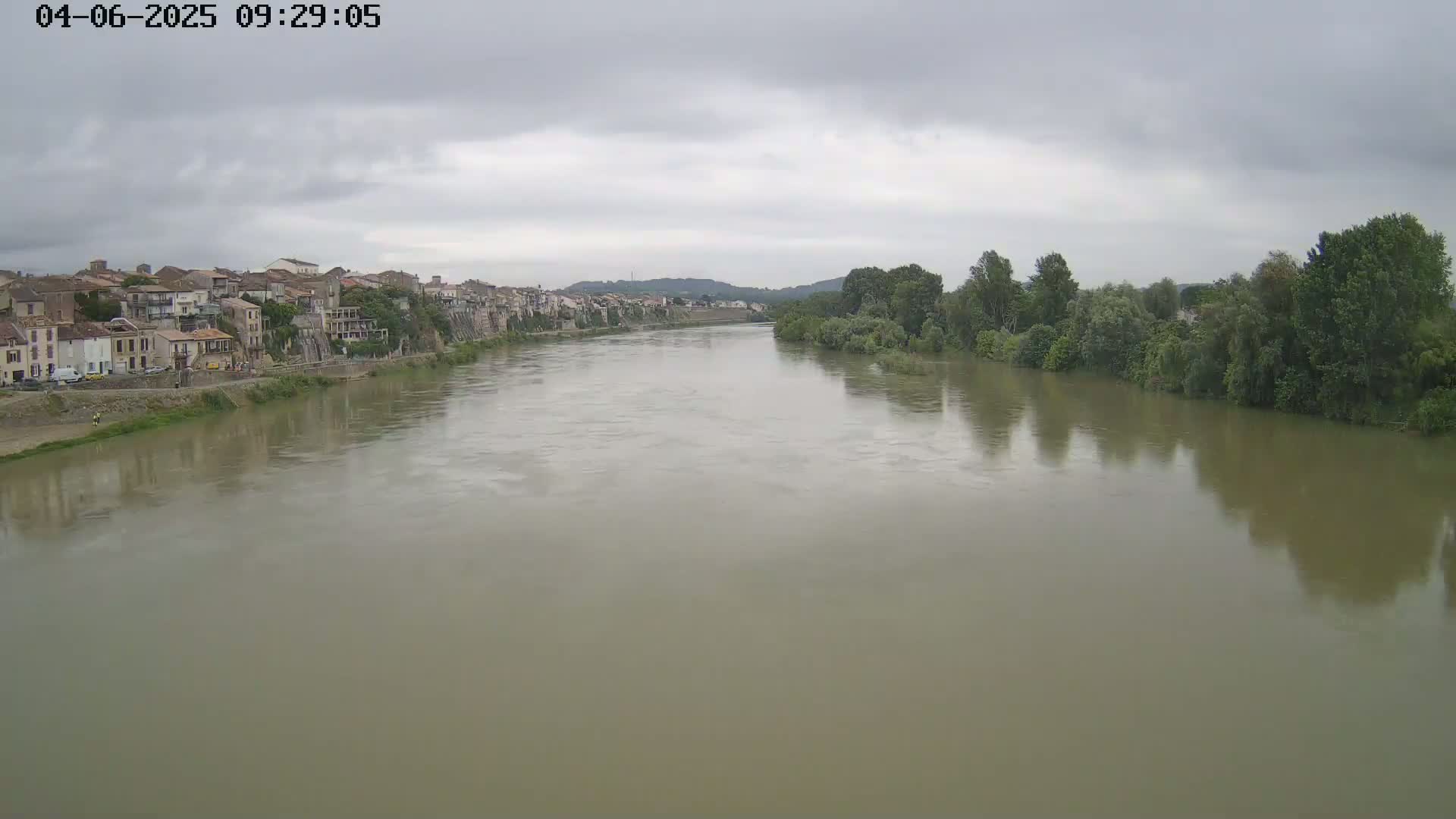 A wide, muddy river flows under an overcast sky past a town of closely built houses and lush green trees.