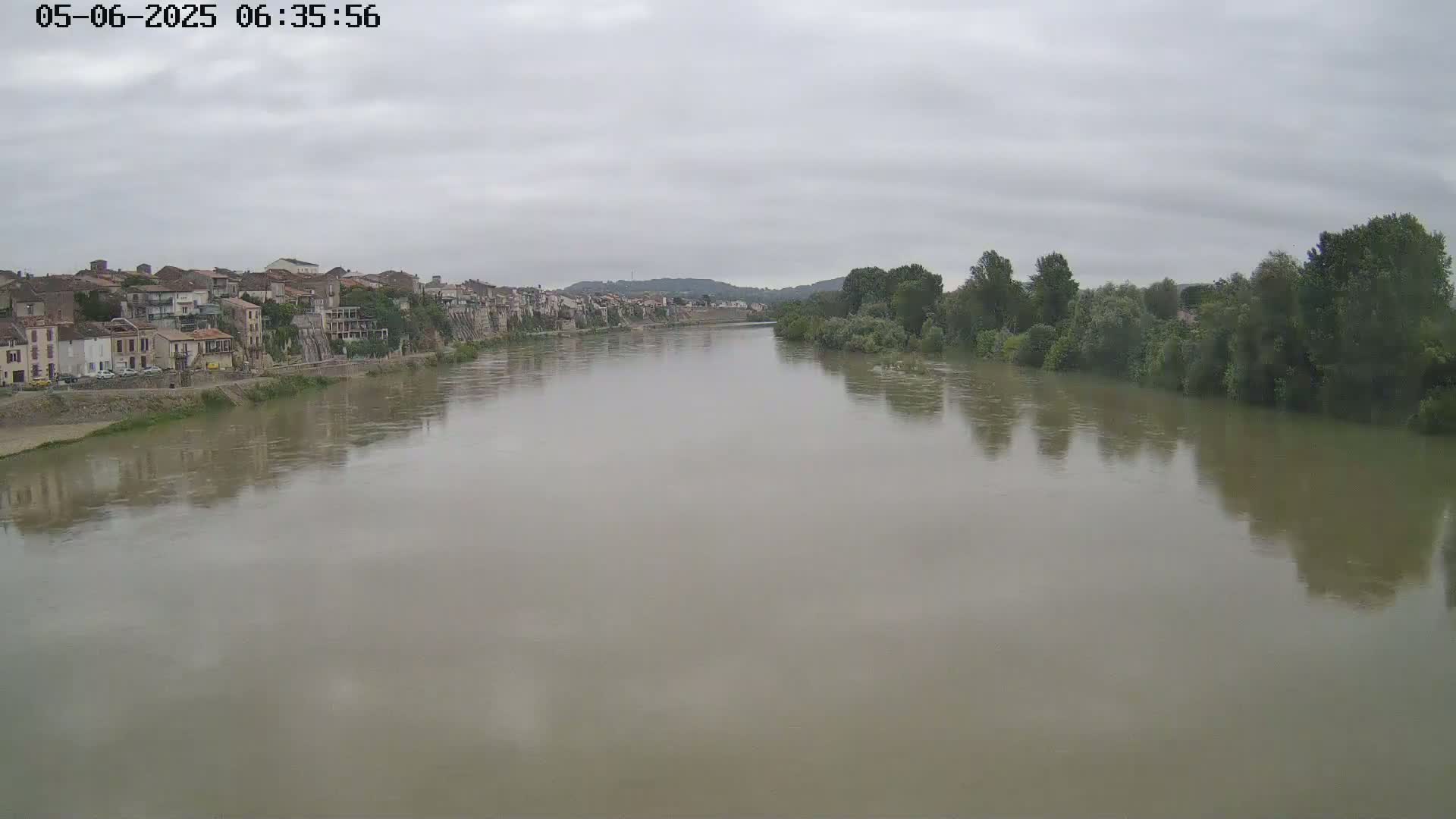 A wide, muddy river flows under an overcast sky past a town on one bank and trees on the other.
