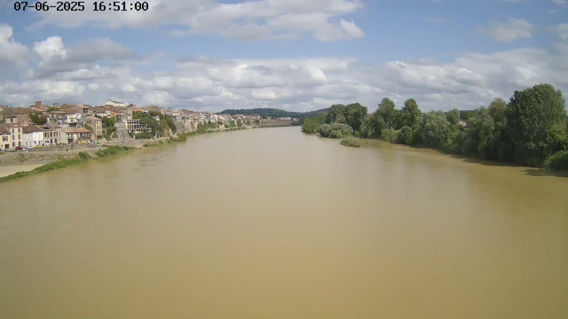 A wide, muddy river flows past a town under a partly cloudy sky.