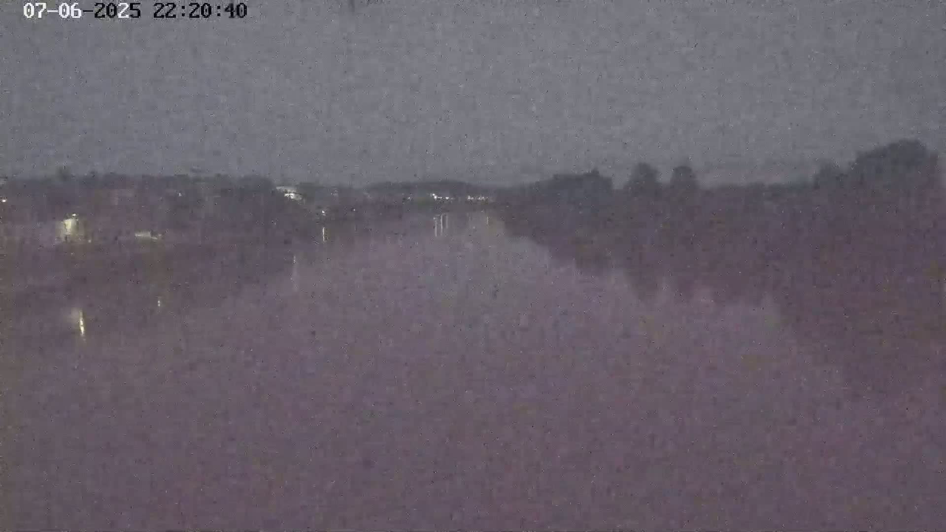 A nighttime long shot of a calm body of water reflecting the lights of a distant town under a dark, overcast sky.