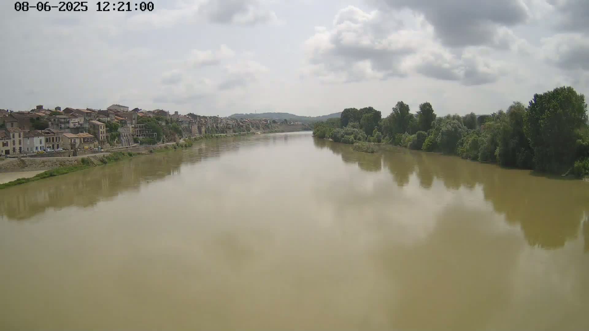 A wide, muddy river flows past a town on one bank and a line of trees on the other under a partly cloudy sky.