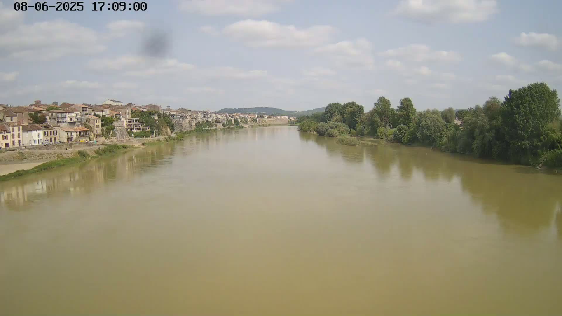 A wide, muddy river flows past a town of low buildings under a partly cloudy sky.