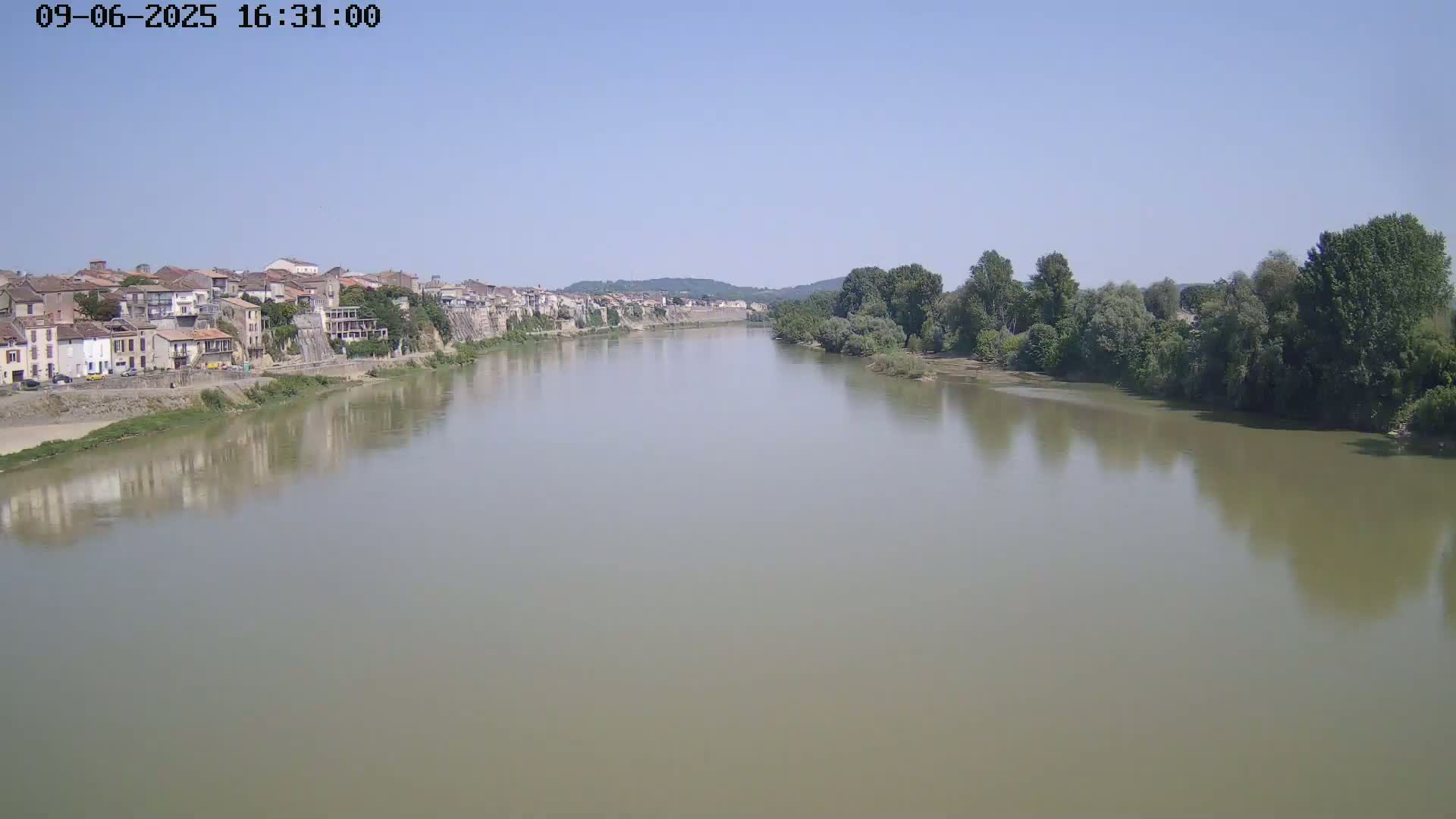 A wide, calm river flows past a town nestled on a hillside under a clear sky.