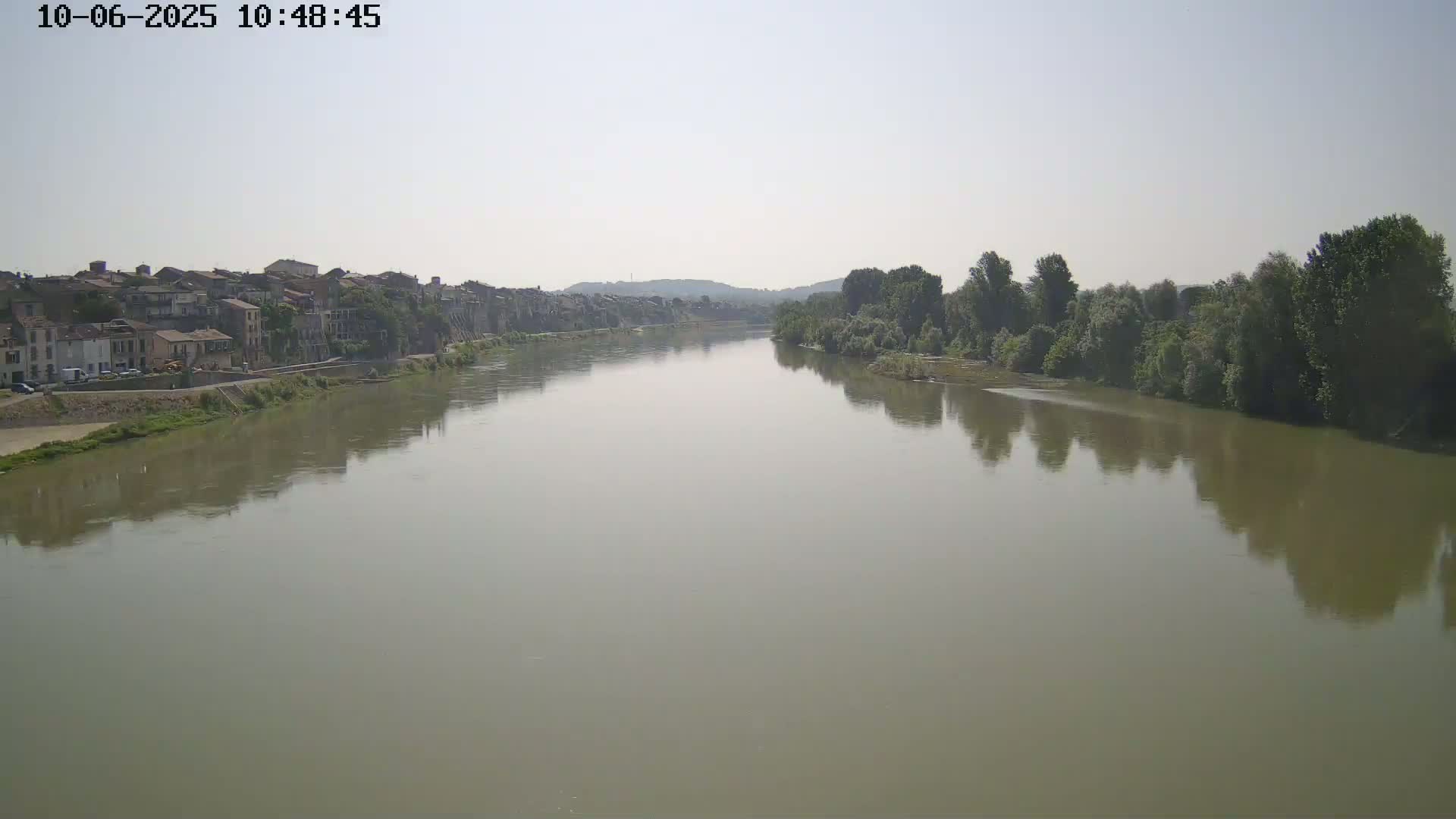 A wide, calm river reflects the hazy sky and the buildings and trees lining its banks.