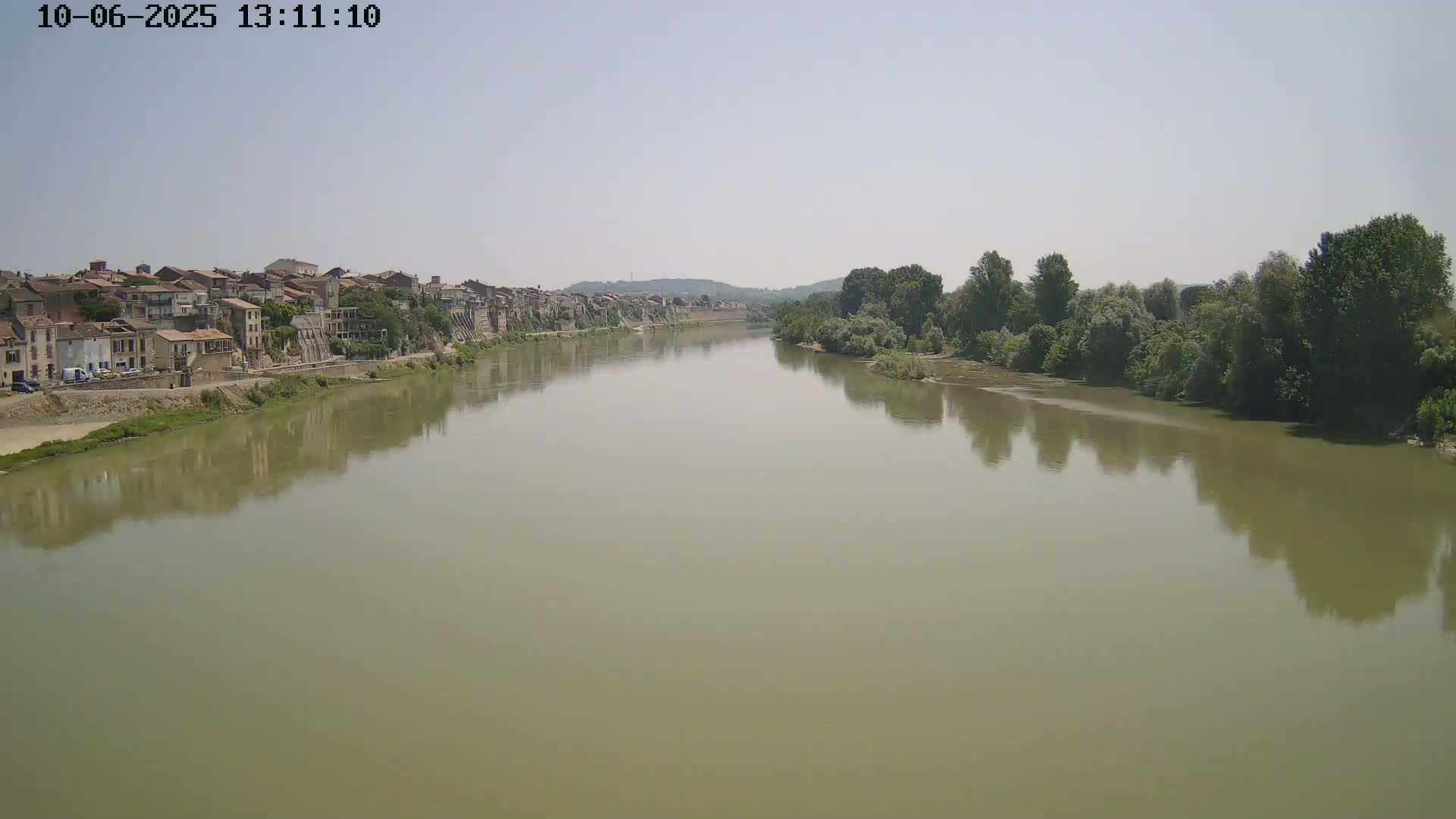 A wide, muddy river reflects the hazy sky and a line of trees and buildings on its banks.