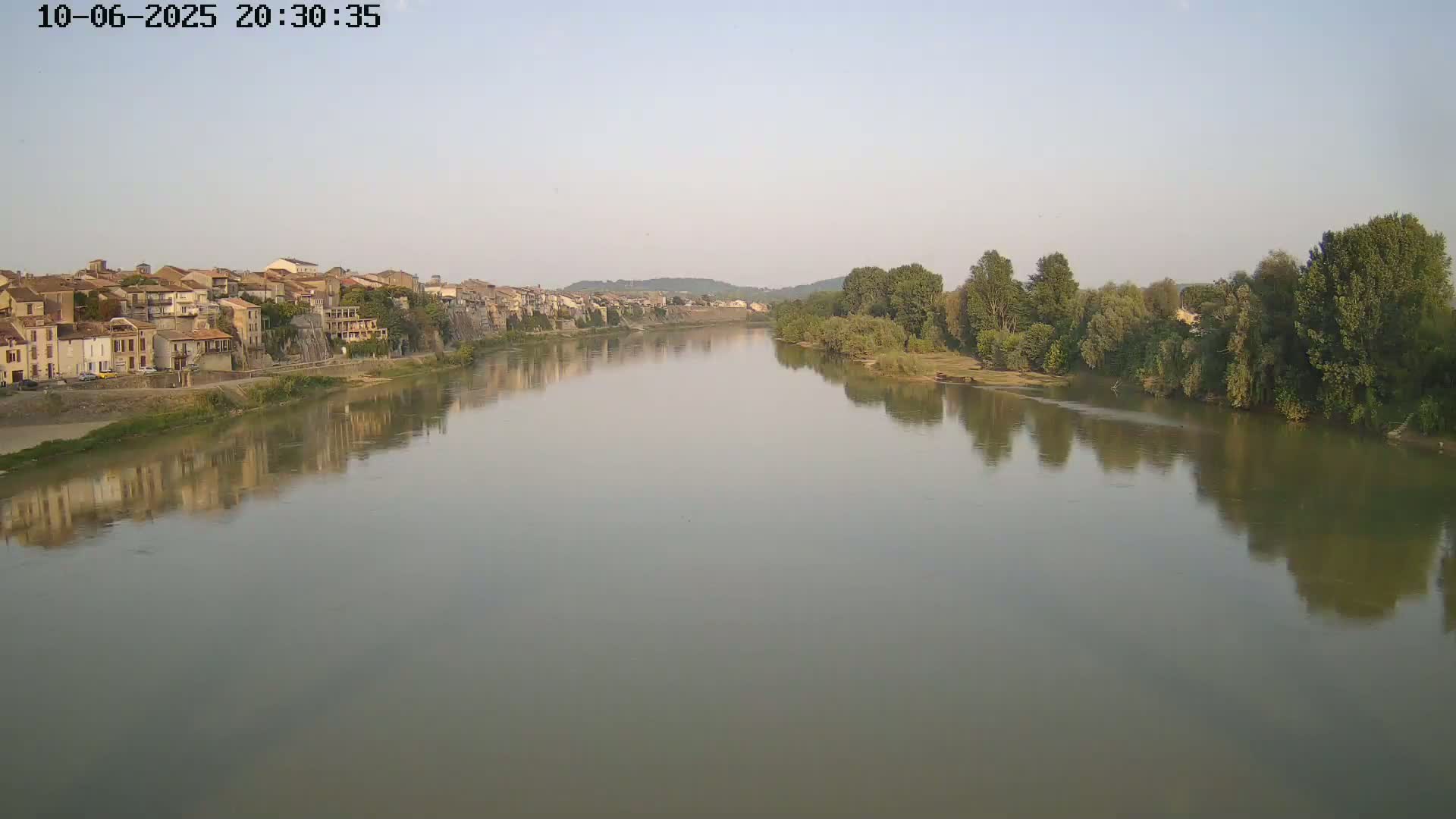 A calm river reflects a town and tree-lined bank under a hazy sky.