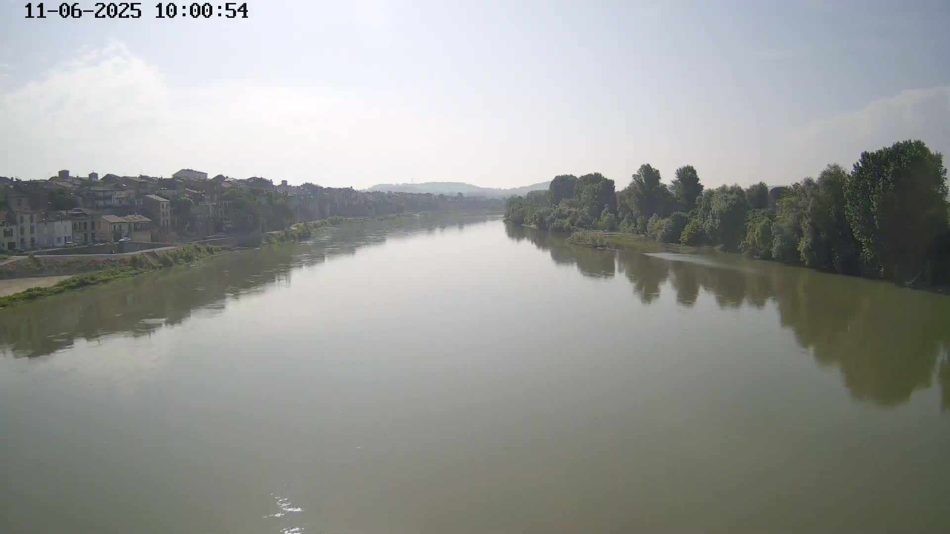 A wide, calm river reflects a hazy sky and the greenery of its banks, with a town visible on one side.