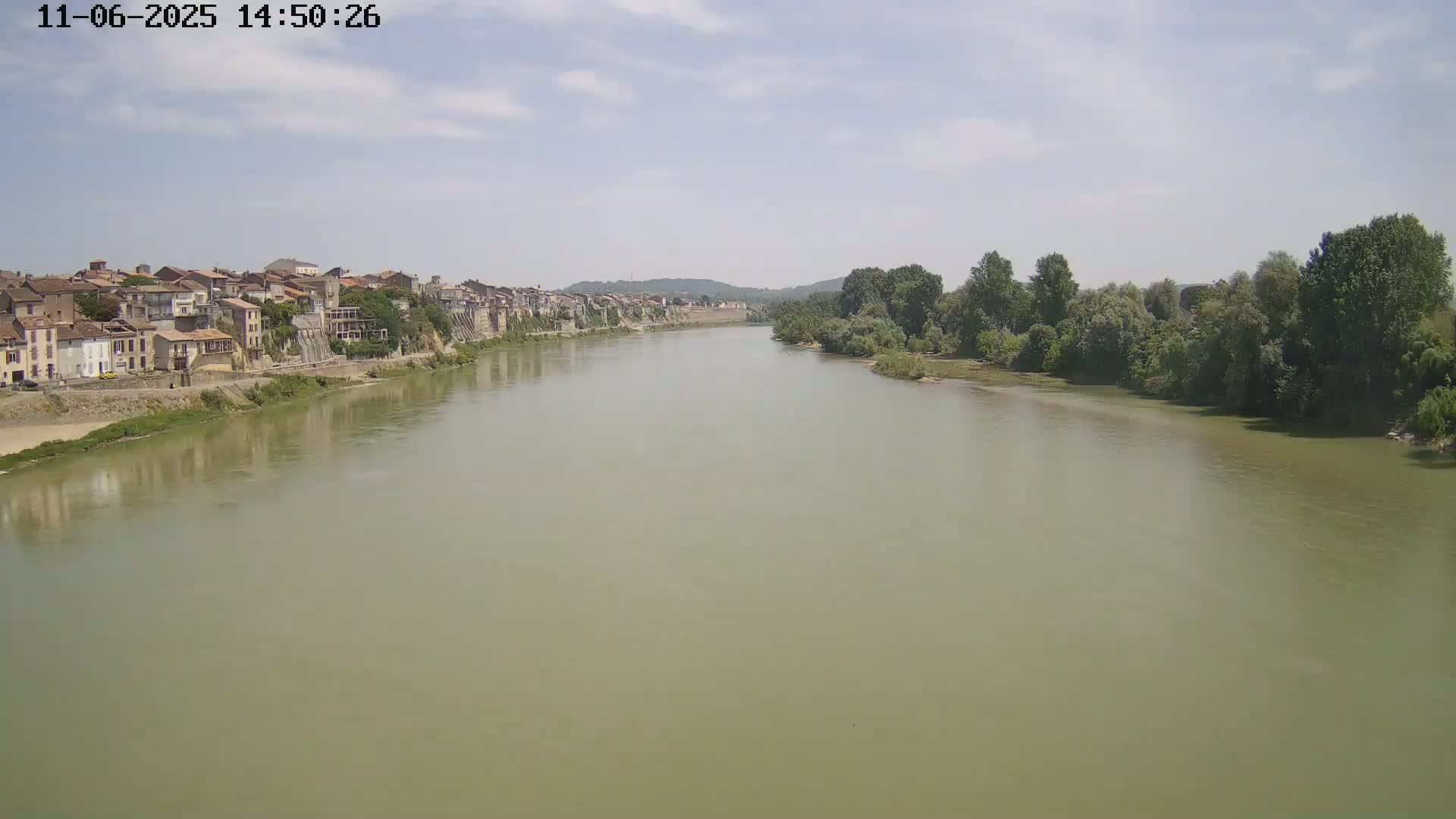 A wide, murky river flows past a town of buildings on a partly cloudy day.
