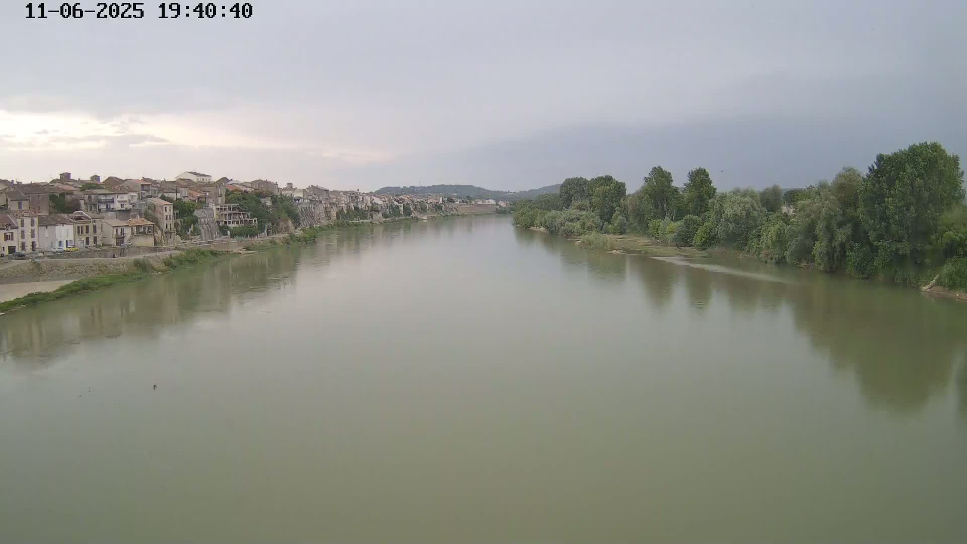 A wide, calm river flows past a town of closely clustered buildings under an overcast sky.