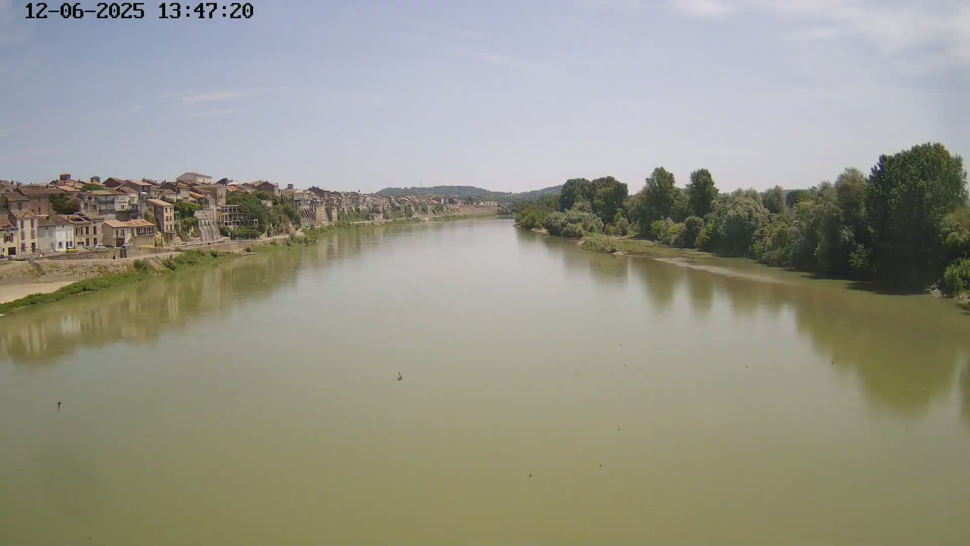 A wide, calm river flows past a town of old buildings under a mostly clear sky.