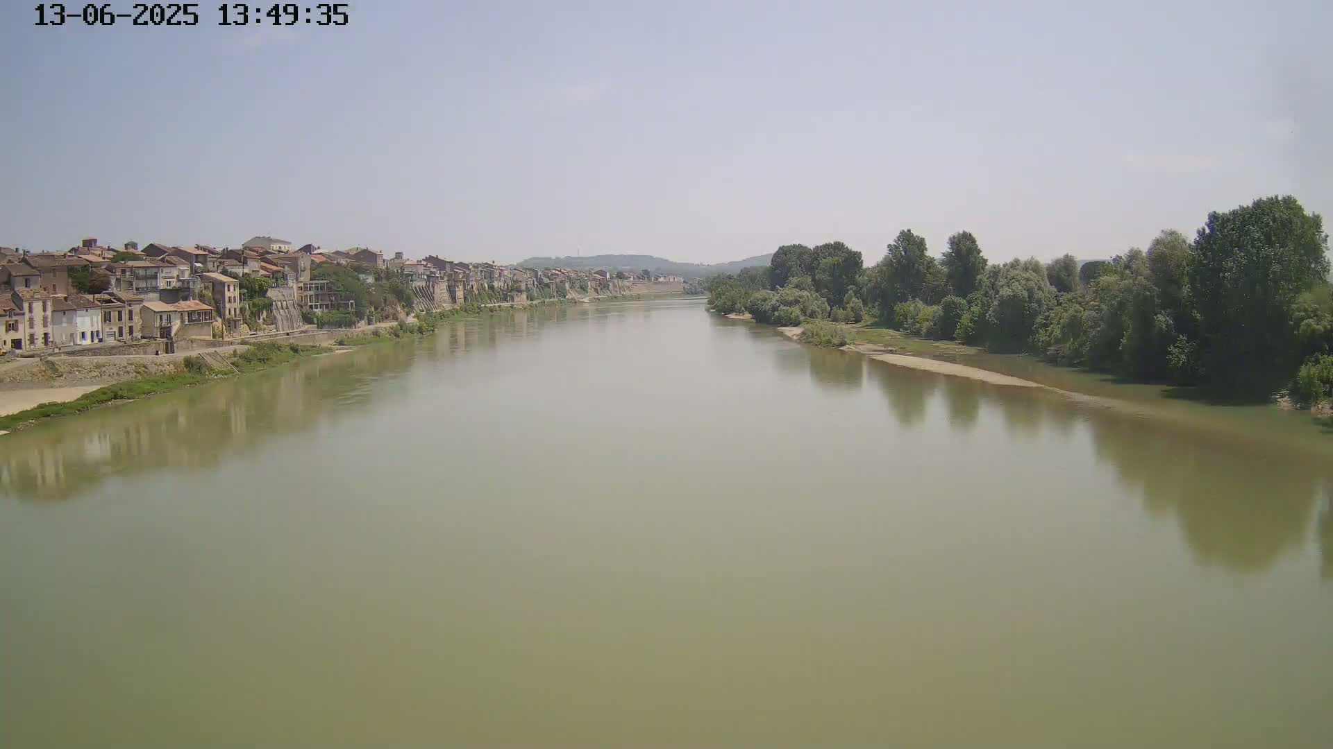 A wide, murky river flows past a town of closely situated buildings under a hazy sky.